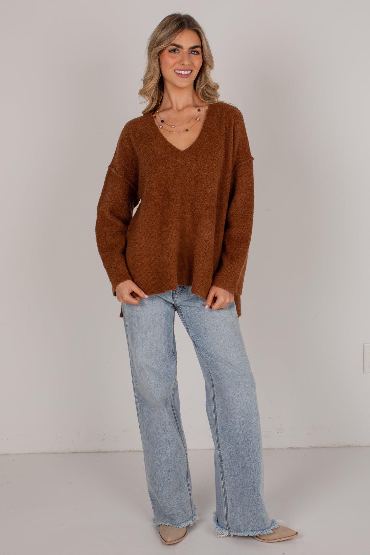 A woman modeling an oversized brown knit sweater with a V-neck, paired with light-wash wide-leg jeans and beige flats, standing against a plain off-white studio wall.