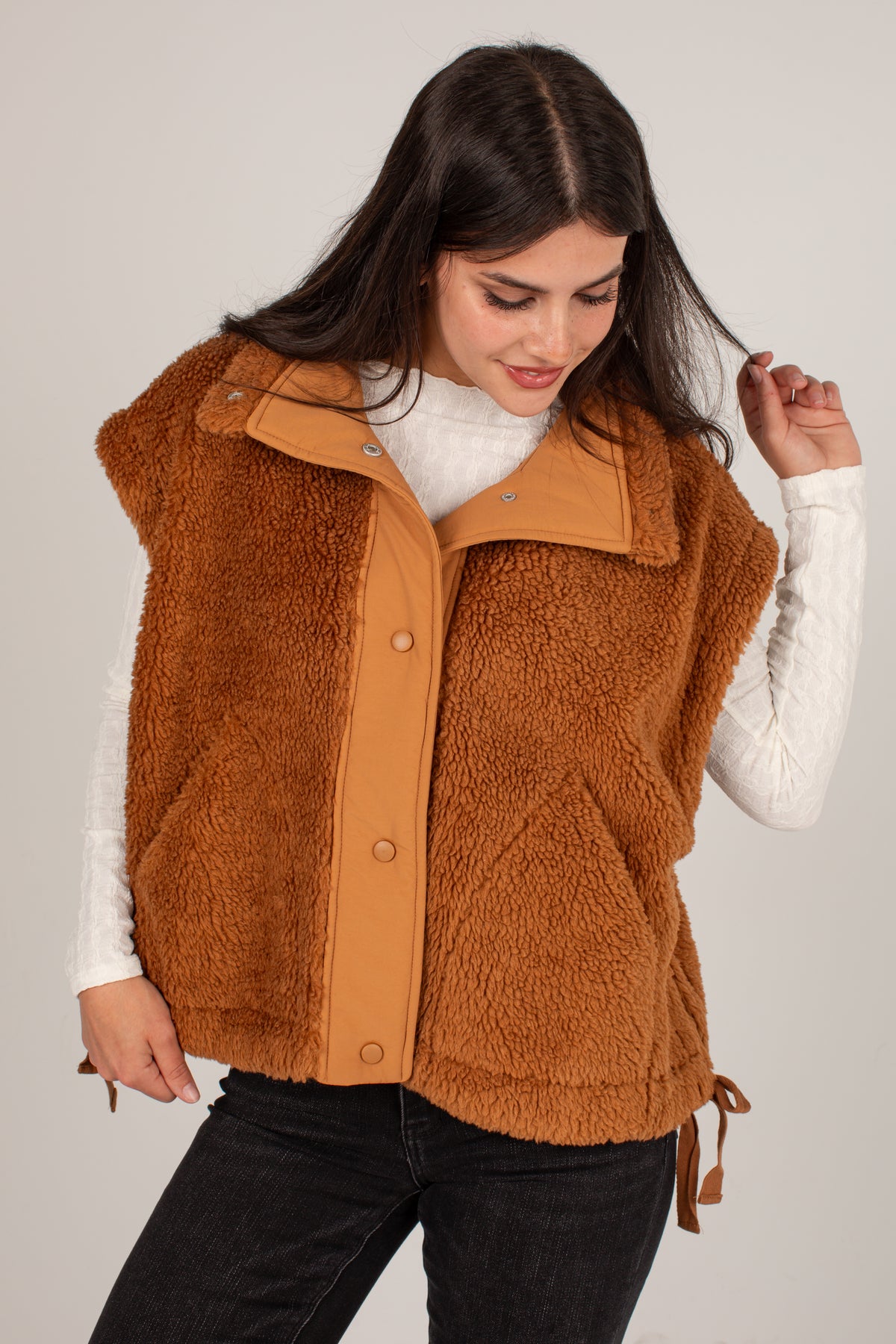 A woman wearing a brown sherpa fleece vest with tan leather trim over a white long-sleeve top and black pants, photographed against a light gray studio background.