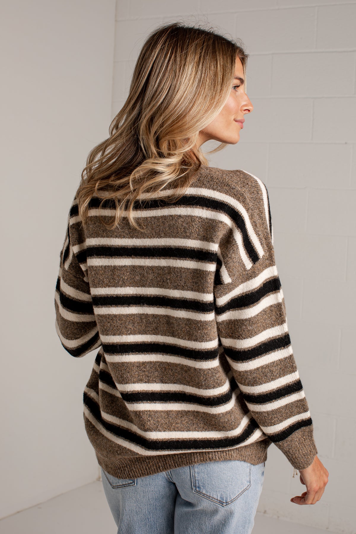 A woman wearing a brown, black, and cream striped knit sweater, shown from a three-quarters back view against a light gray brick wall.