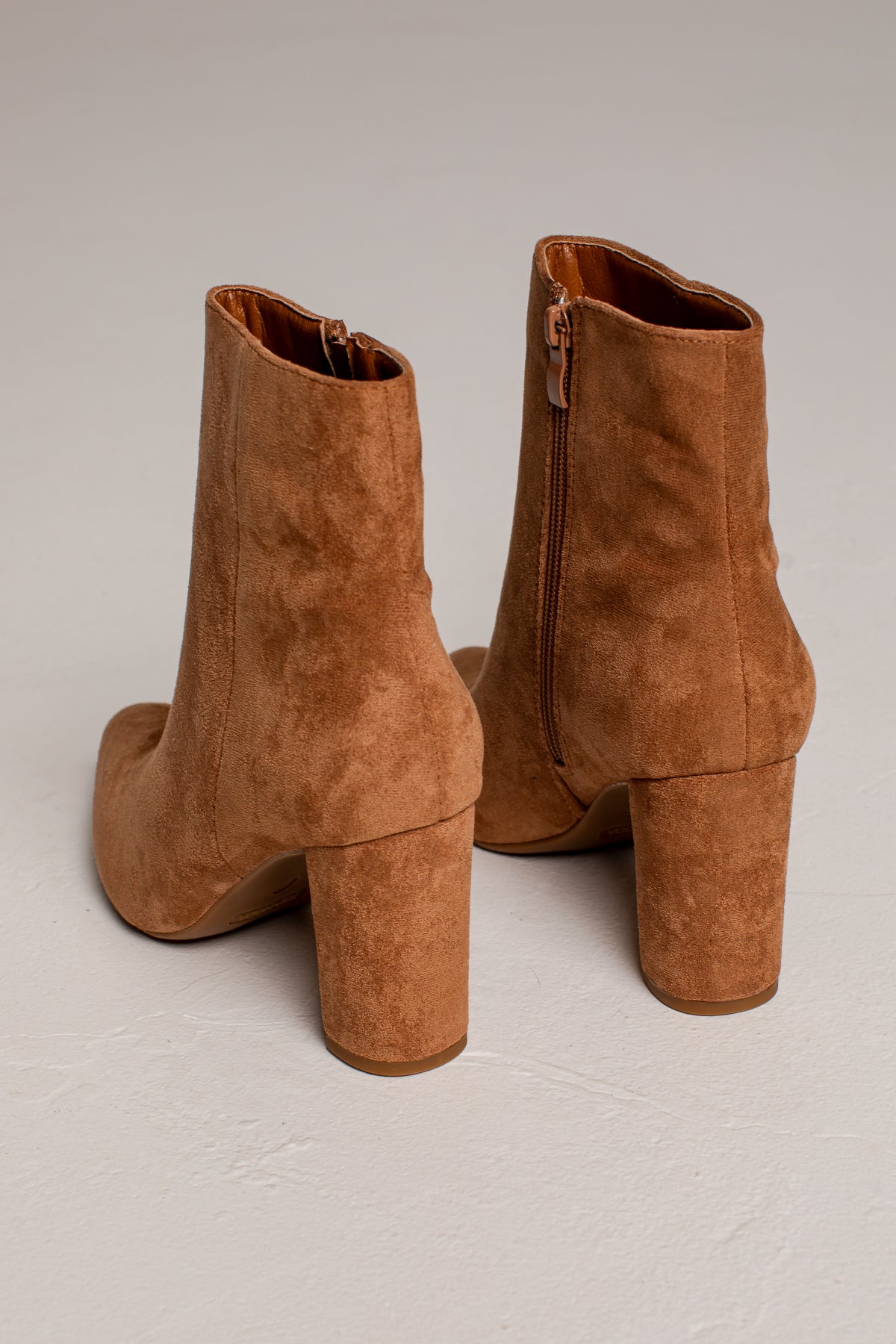 A pair of brown suede ankle boots with block heels, shown from the back on a light gray textured background.
