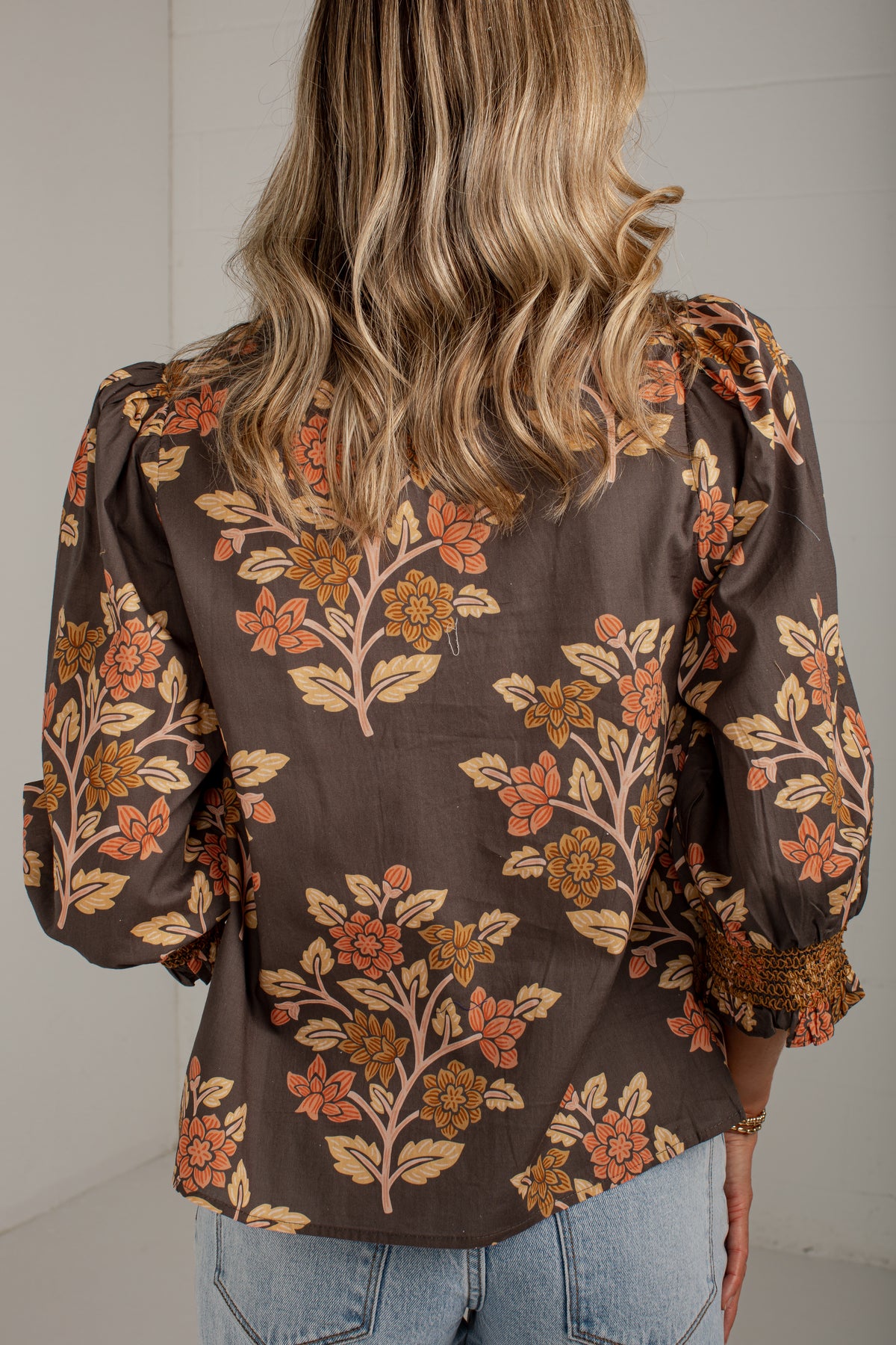 A woman viewed from the back wearing a brown long-sleeve blouse with a floral pattern in orange, beige, and muted pink, paired with light blue jeans.