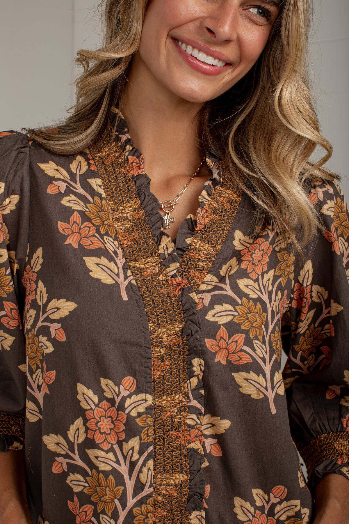 A close-up of a woman wearing a brown blouse with a floral print in orange, beige, and brown, featuring a ruffled V-neckline and embroidered trim.