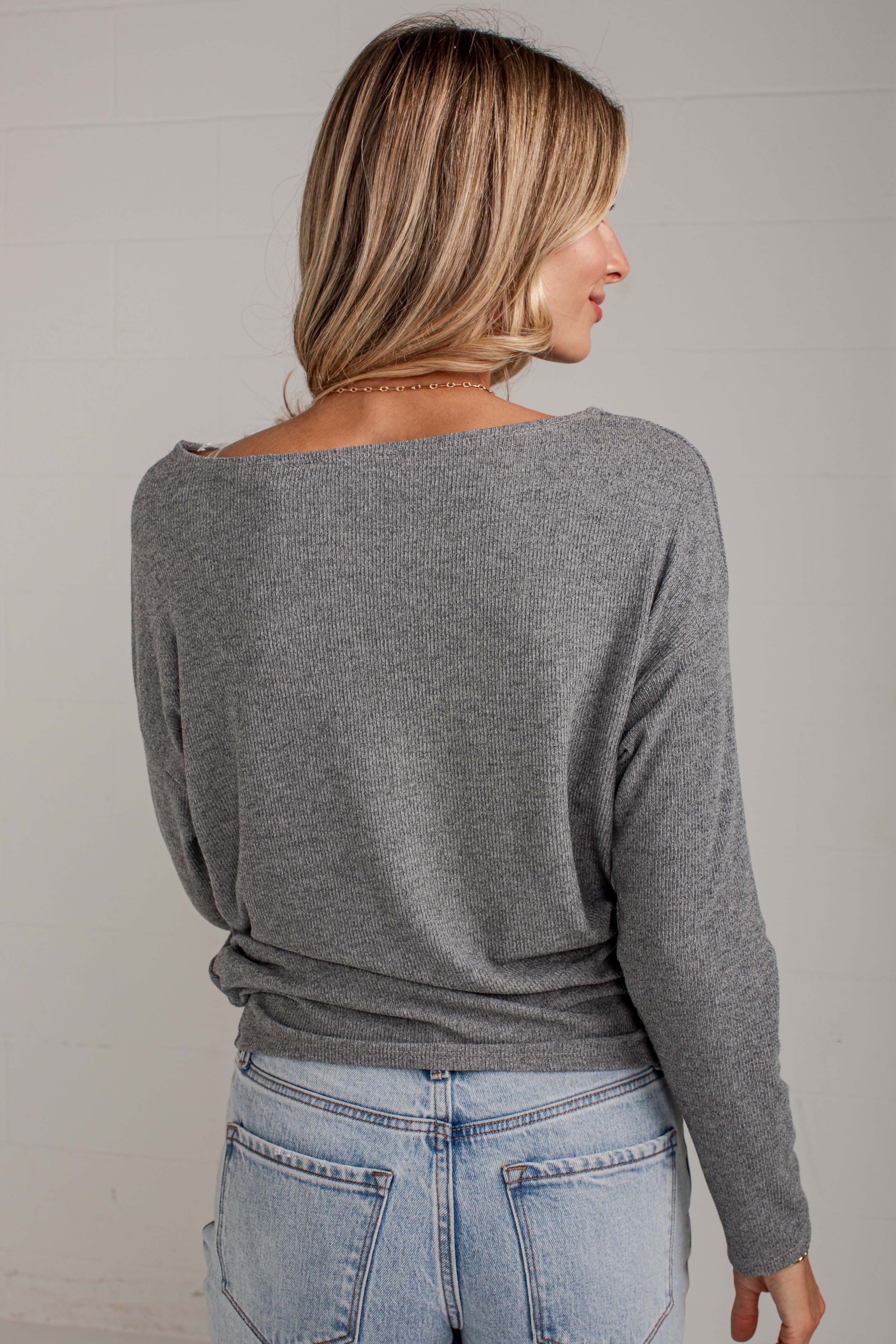 A woman shown from the back wearing a heather gray long-sleeve knit top with a wide boat neckline, paired with light blue jeans, against a light gray wall.