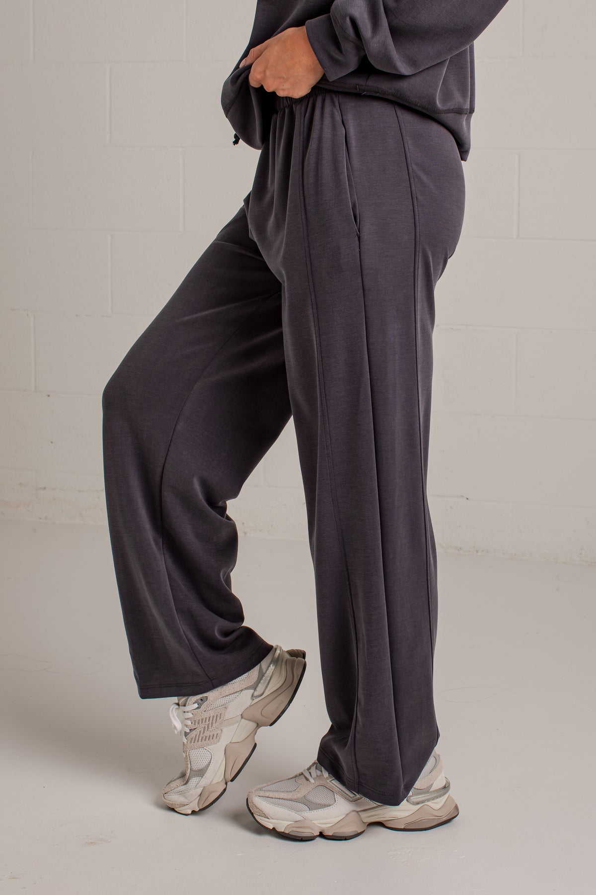 A person wearing loose, wide-leg dark charcoal sweatpants and chunky off-white sneakers, standing against a light gray brick wall.