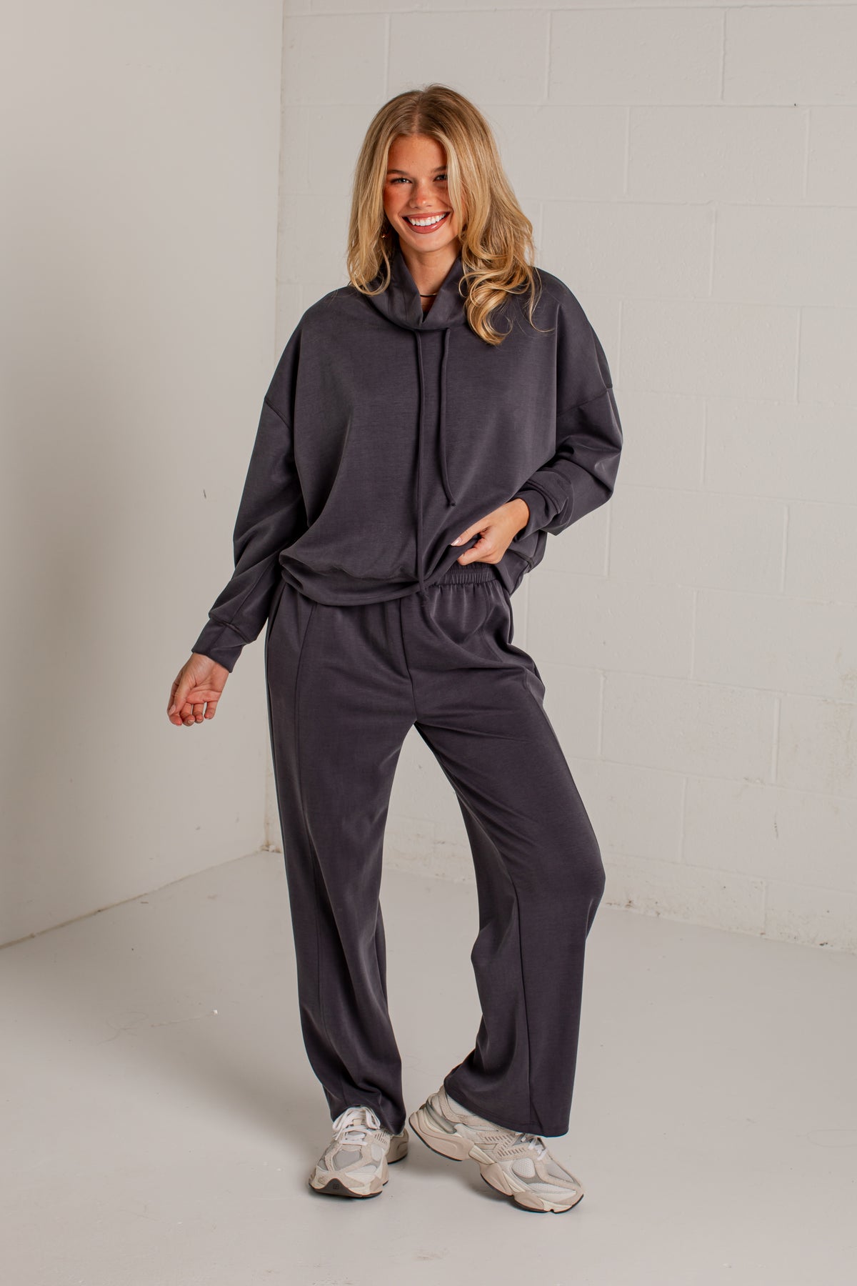 A woman modeling a dark charcoal gray two-piece loungewear set consisting of a hooded sweatshirt and matching wide-leg sweatpants, paired with white sneakers, in a bright studio setting.