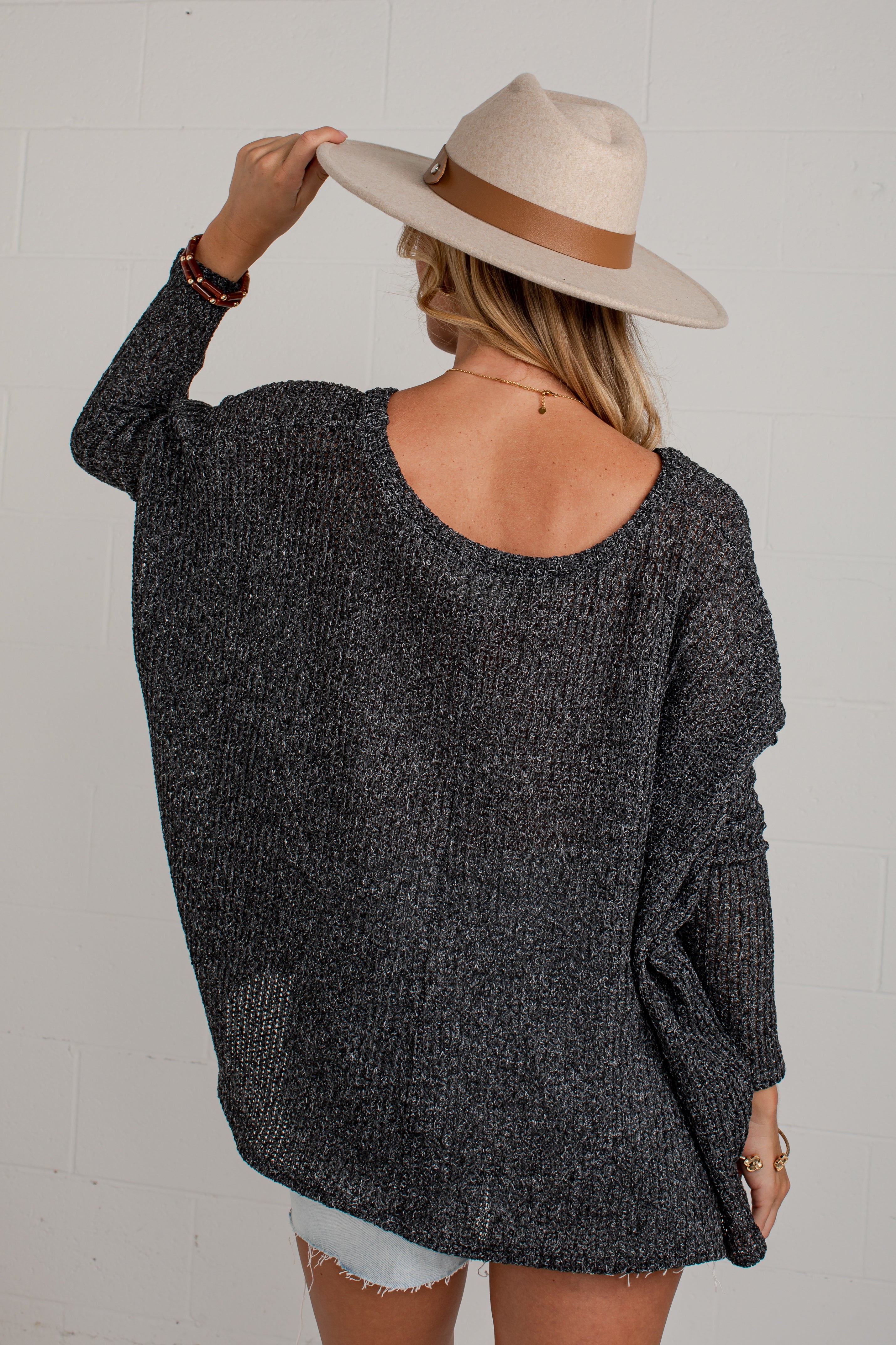 A woman shown from behind wearing a dark gray marled knit sweater, a beige wide-brim hat with a brown band, and light denim shorts against a white brick wall.