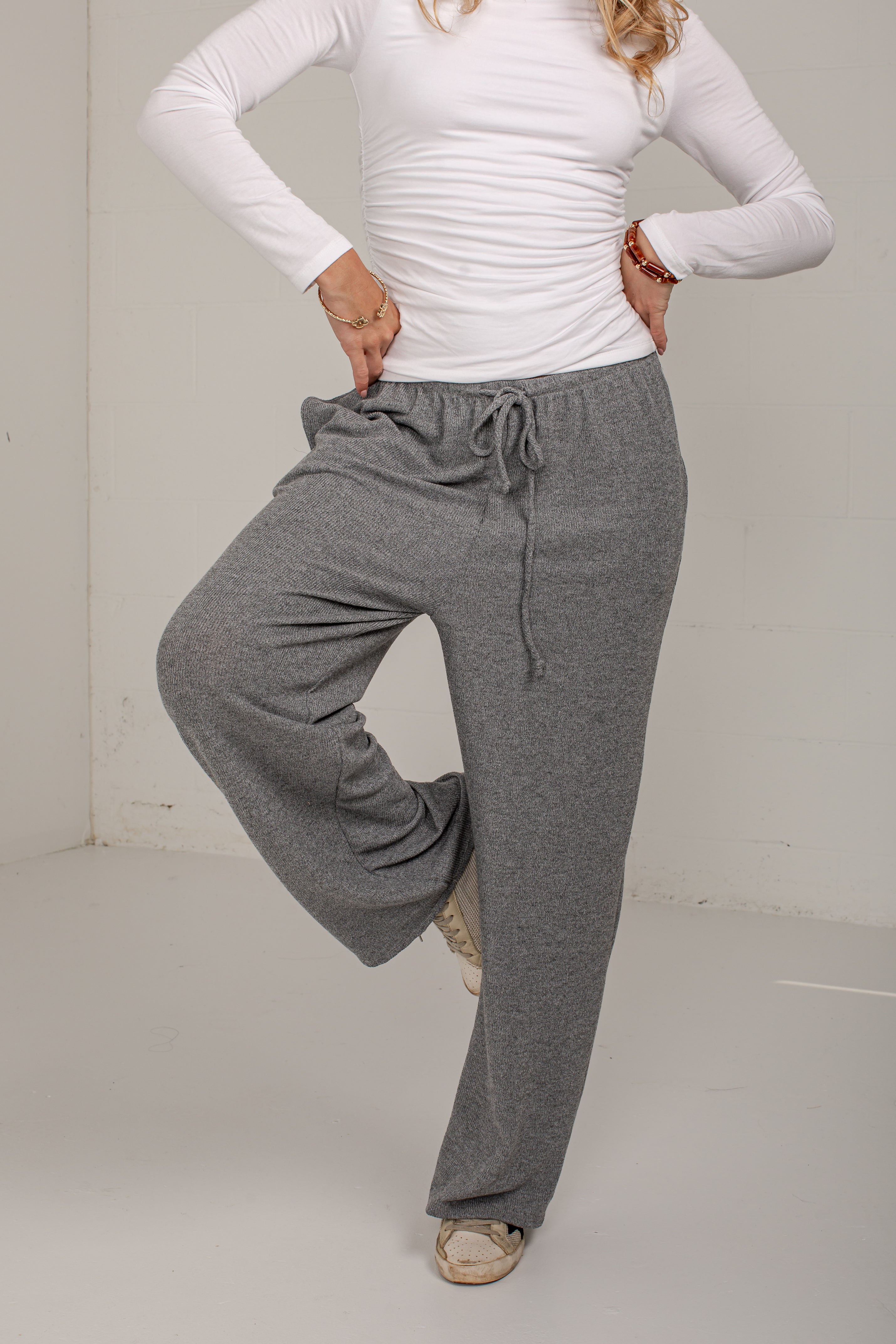 A woman modeling a pair of heather gray wide-leg lounge pants with a drawstring waist, paired with a white long-sleeve top and beige shoes.
