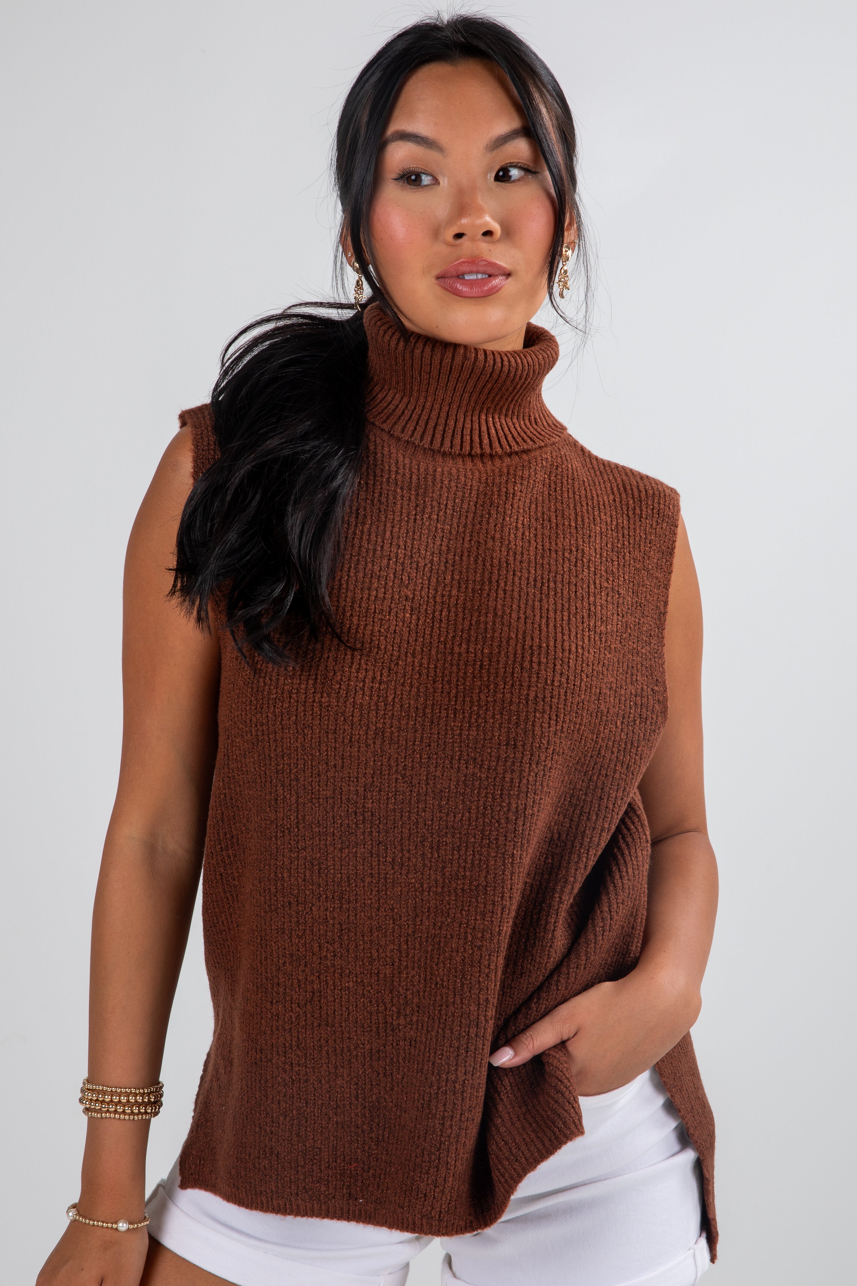 Woman wearing a brown sleeveless sweater over a white top against a plain background