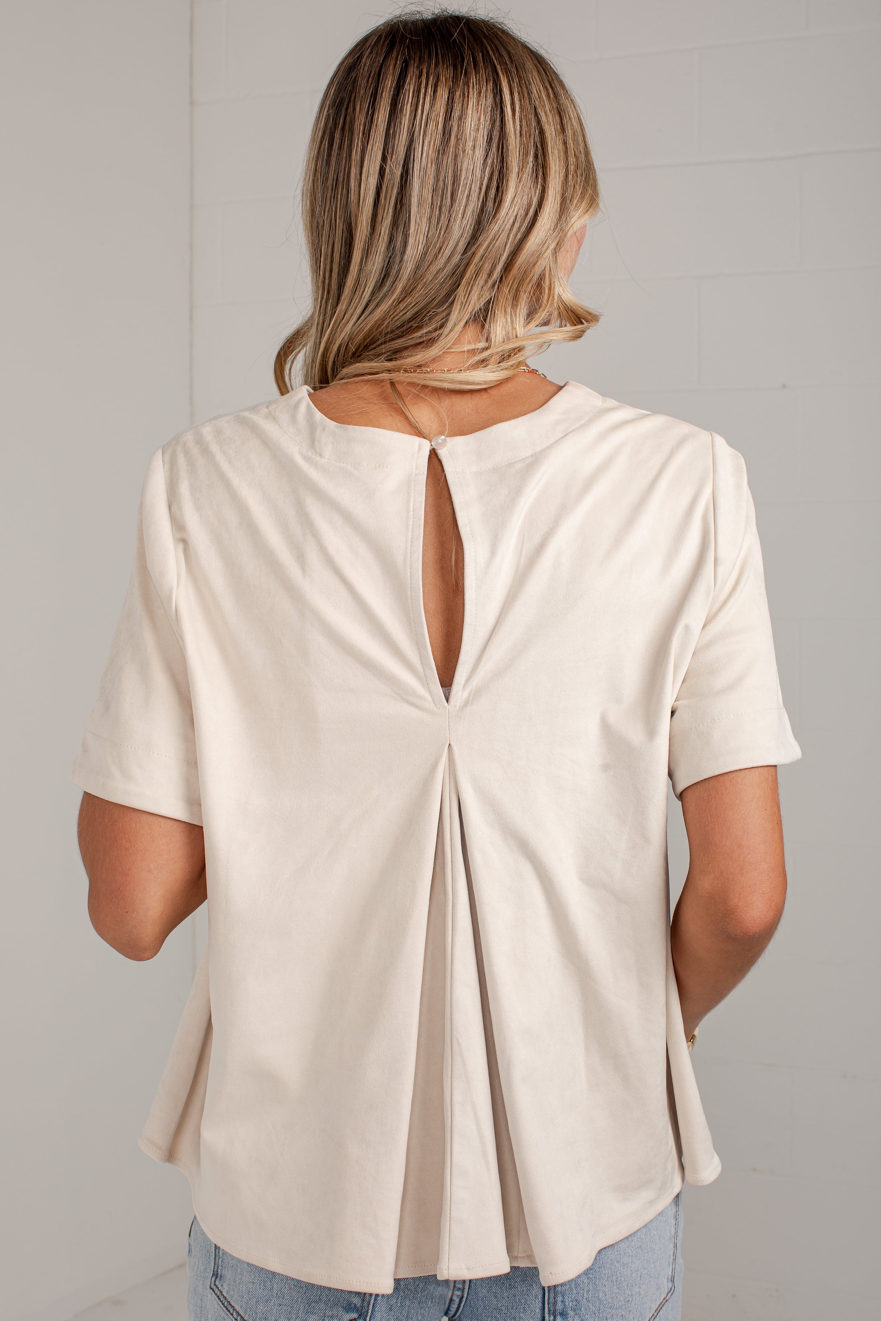 A woman viewed from the back wearing a short-sleeve cream-colored top with a keyhole cutout at the back, paired with light blue jeans against a light gray wall.