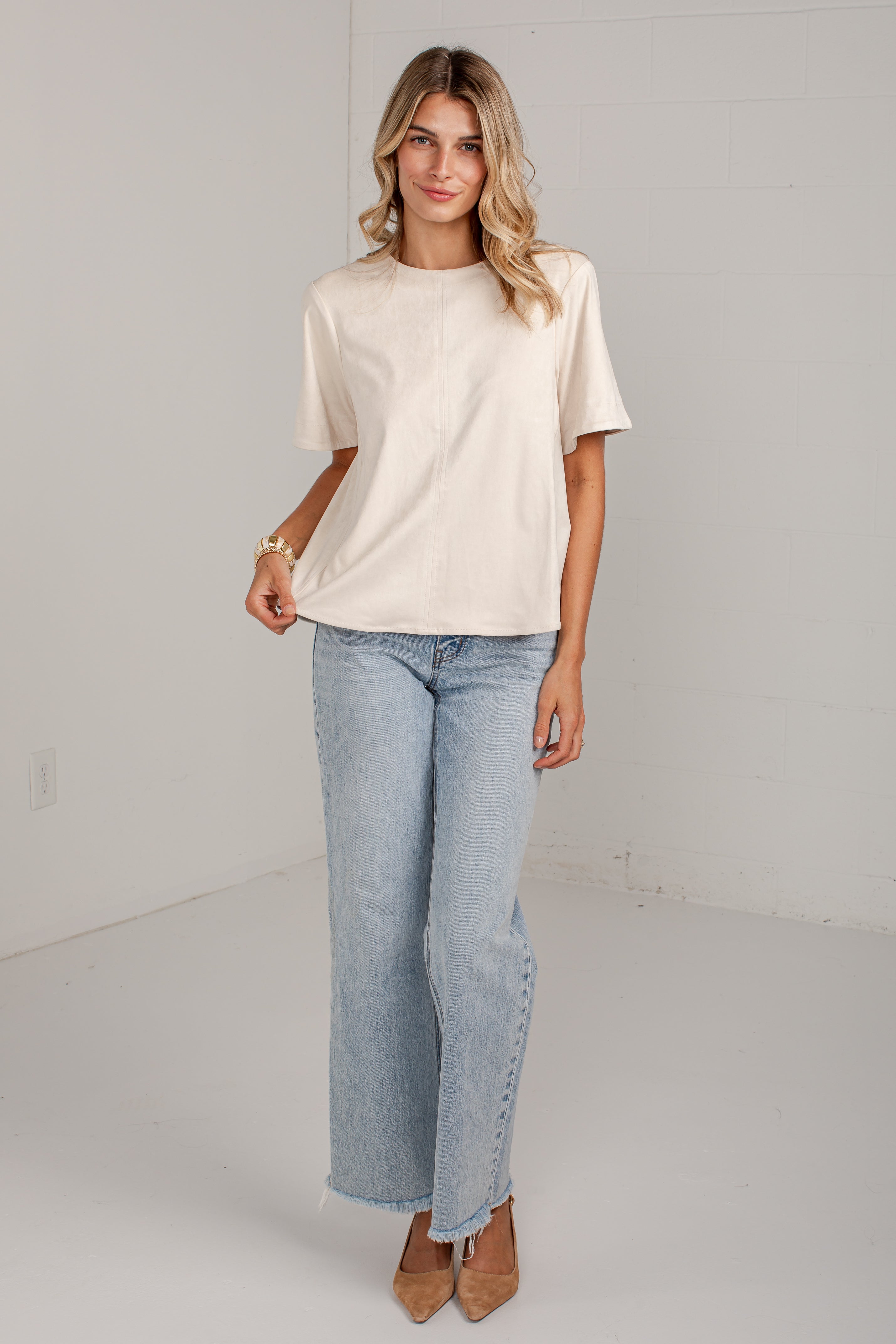 A woman modeling a cream-colored short-sleeve crewneck t-shirt and light-wash wide-leg jeans with side button details, paired with brown pointed-toe heels, in a bright white studio setting.