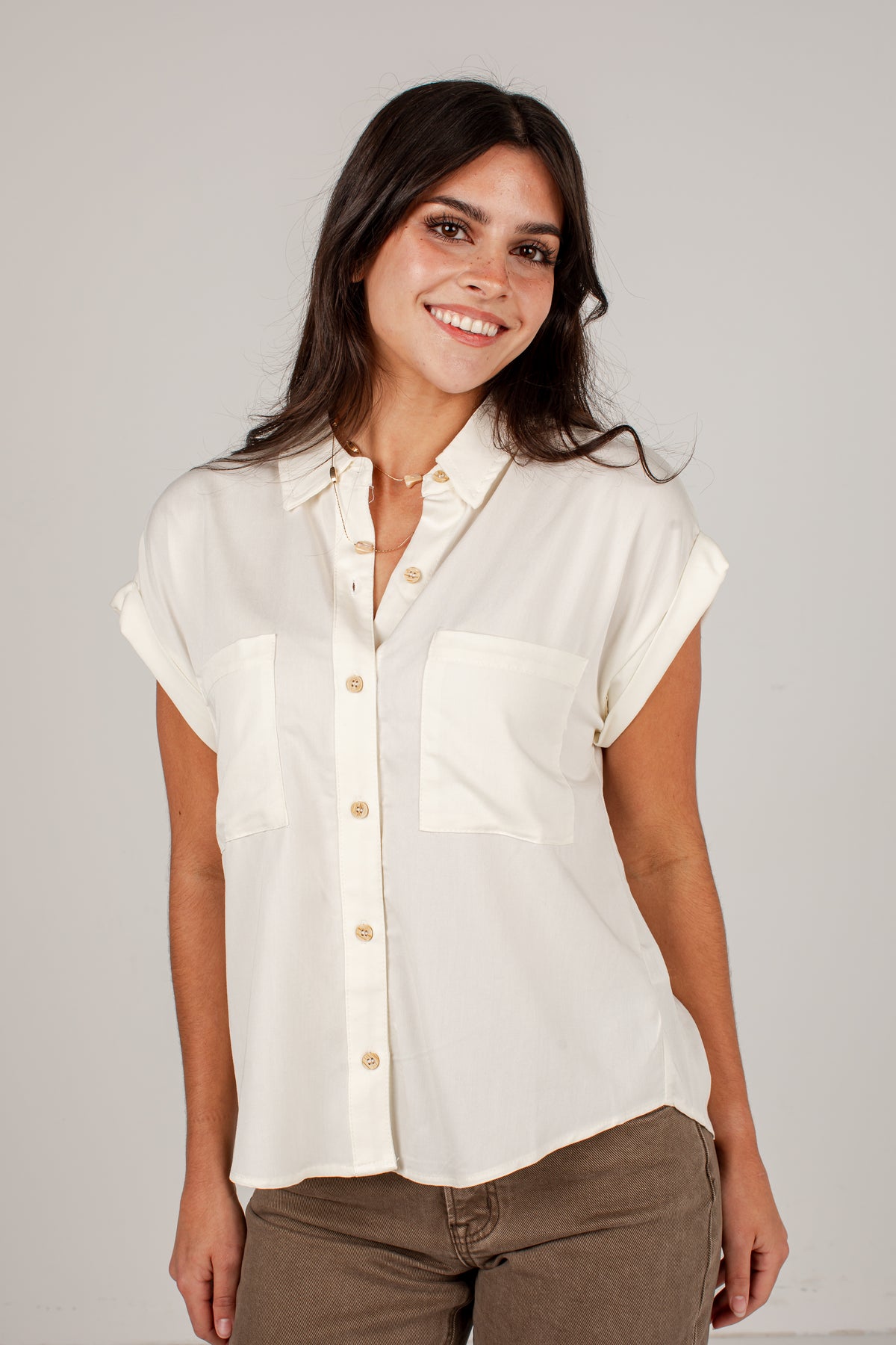 A woman wearing a short-sleeve button-up shirt in off-white with two chest pockets, paired with brown pants, standing against a light gray studio background.