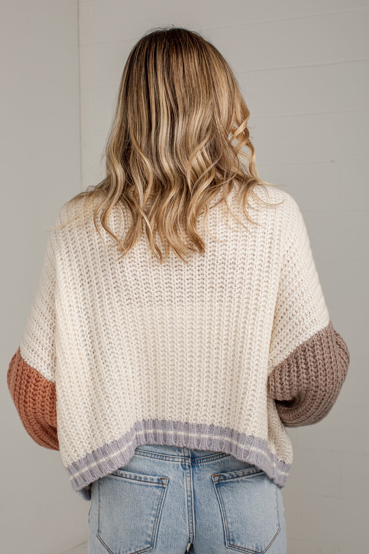 A woman viewed from behind wearing a chunky knit sweater with cream body, terracotta sleeves, and a gray striped hem, paired with light blue jeans.