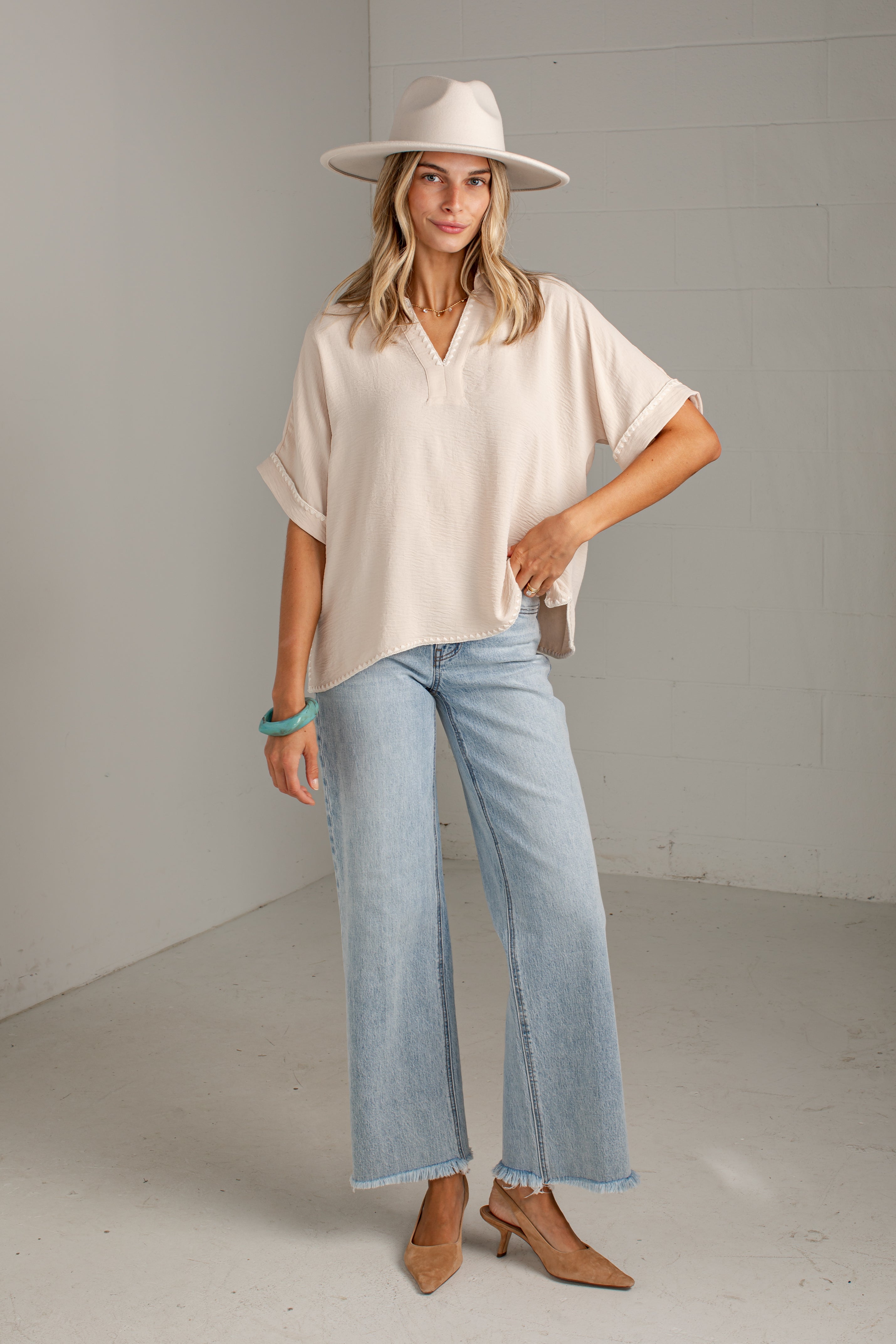 A woman modeling a cream short-sleeve collared blouse, light-wash wide-leg jeans, a cream wide-brim hat, and tan heeled mules in a bright studio setting.