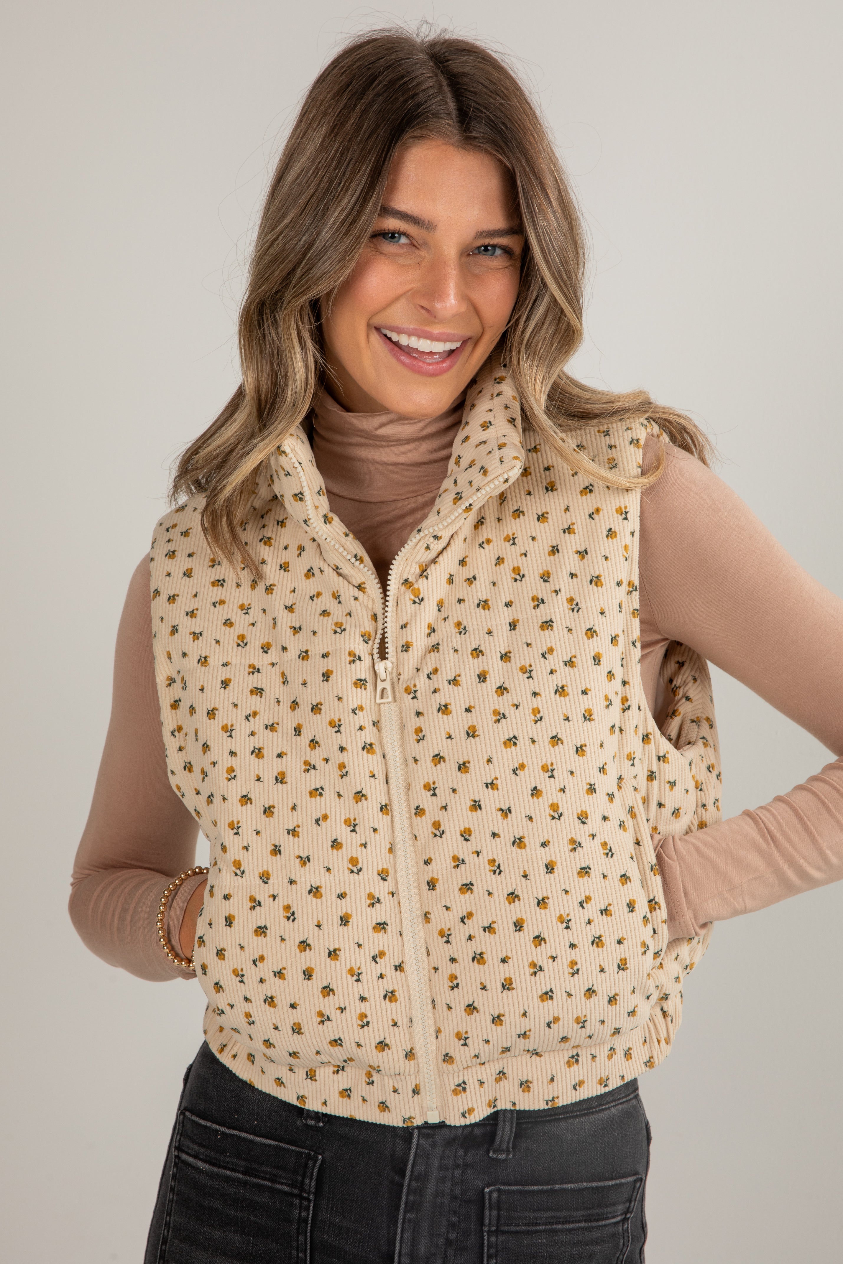 Woman wearing a beige patterned vest over a brown top and black pants on a plain background