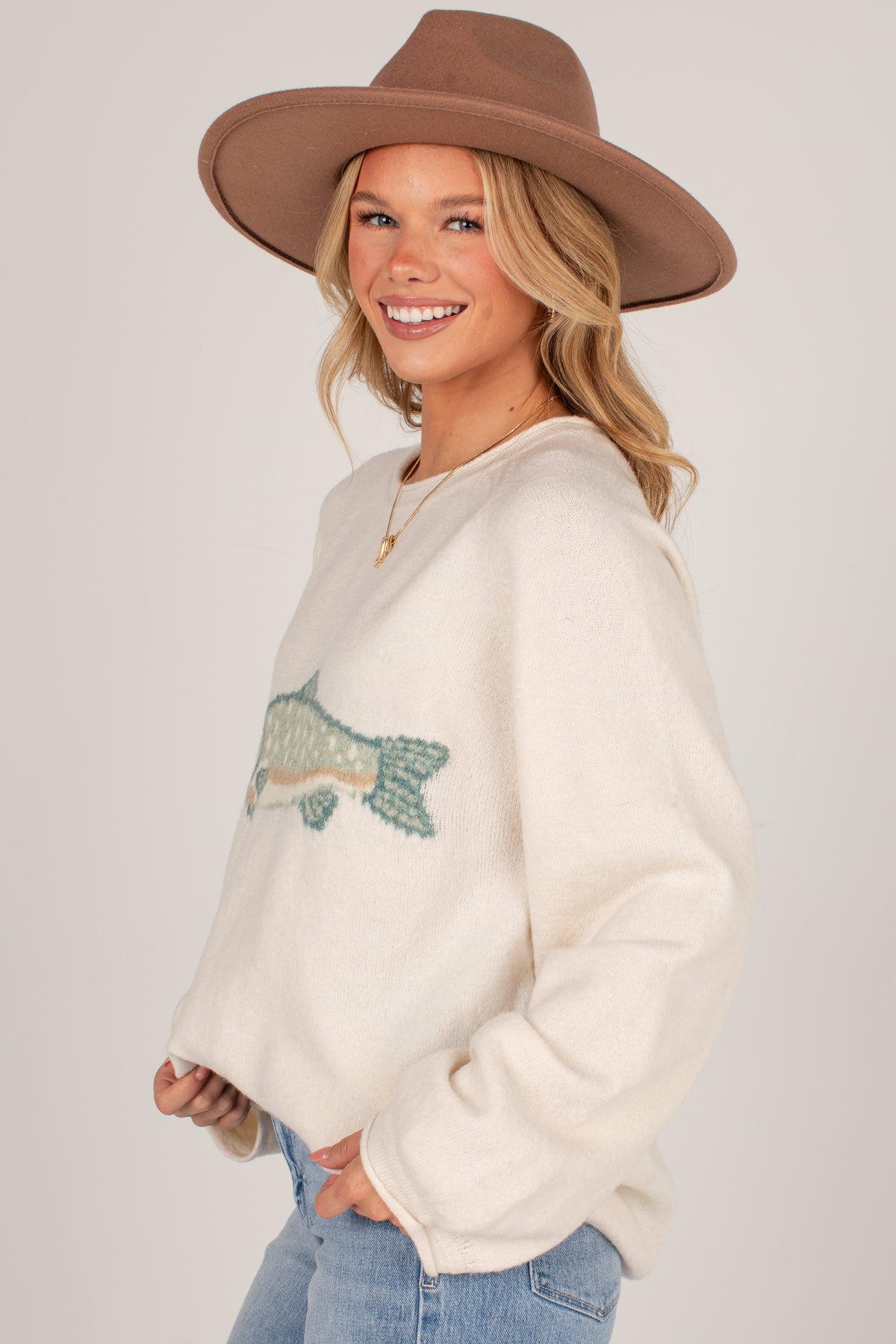 A woman wearing a cream-colored fuzzy sweater with a green and blue fish graphic on the front, paired with a brown wide-brim hat and light blue jeans, photographed against a light gray studio background.