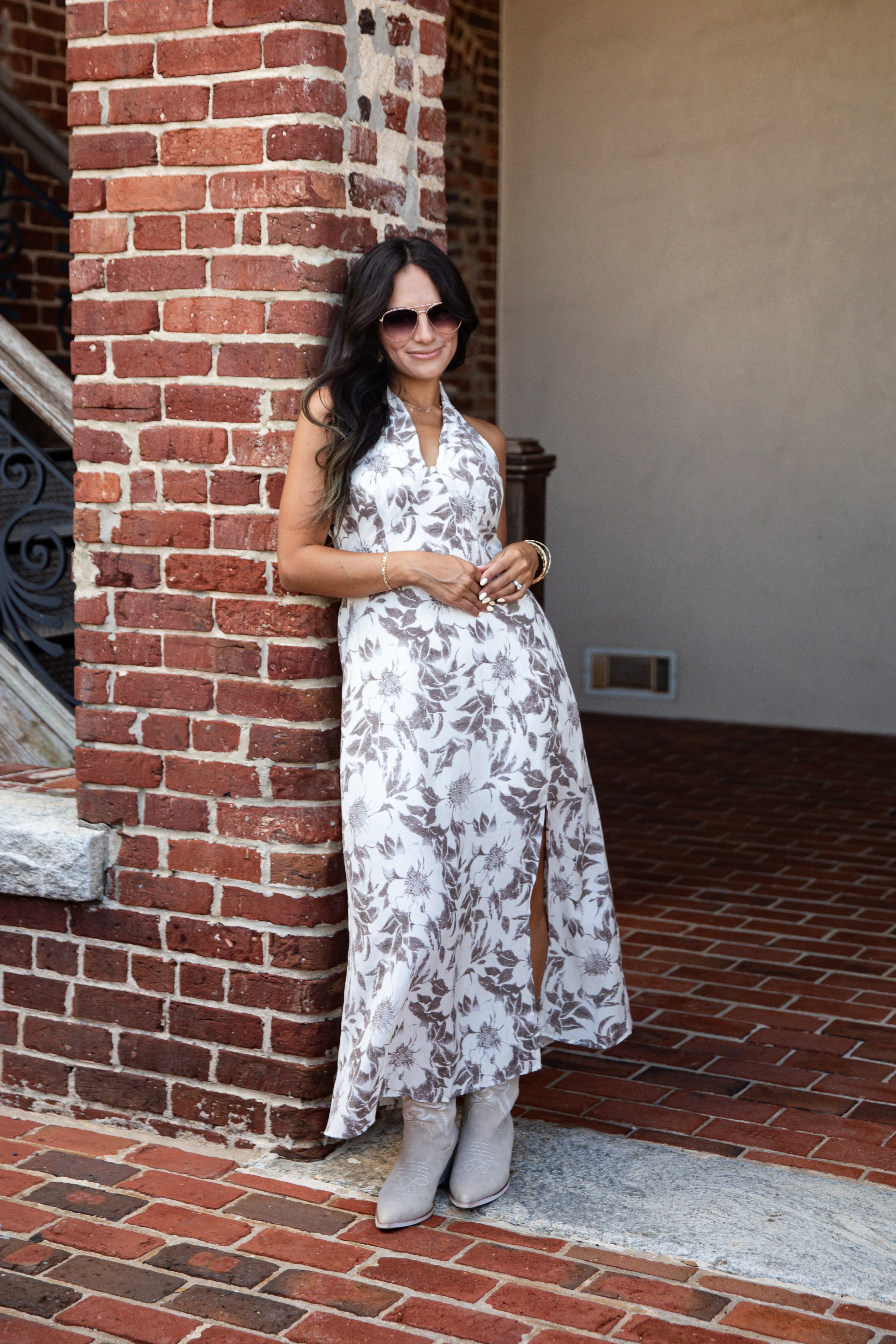 A woman wearing a sleeveless white halter maxi dress with a gray botanical leaf pattern and gray boots, posing outdoors against a red brick column.