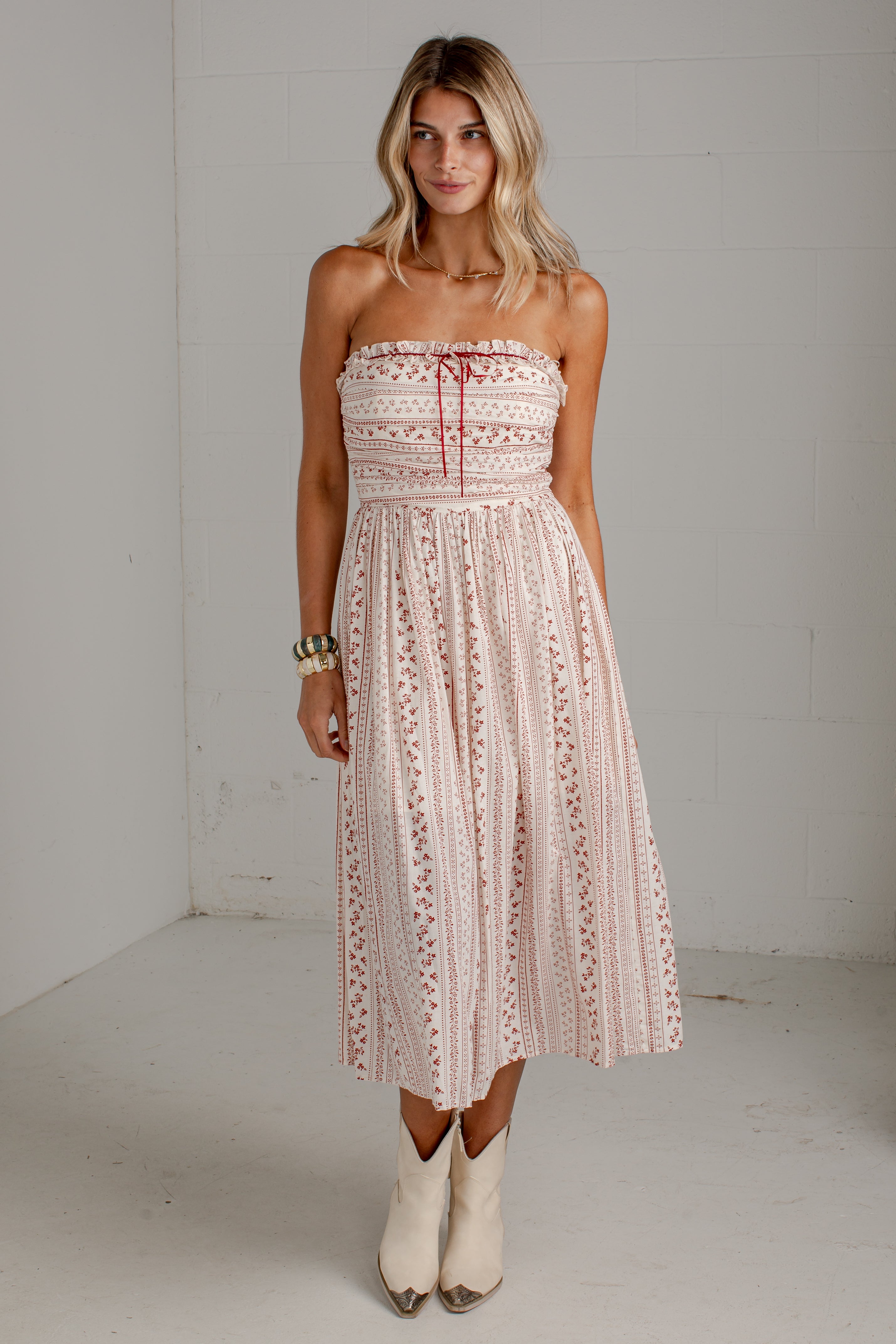 A woman modeling a strapless midi dress with a cream and pink floral pattern, featuring a tie-neck detail and a flared skirt, paired with white cowboy boots against a white brick wall background.