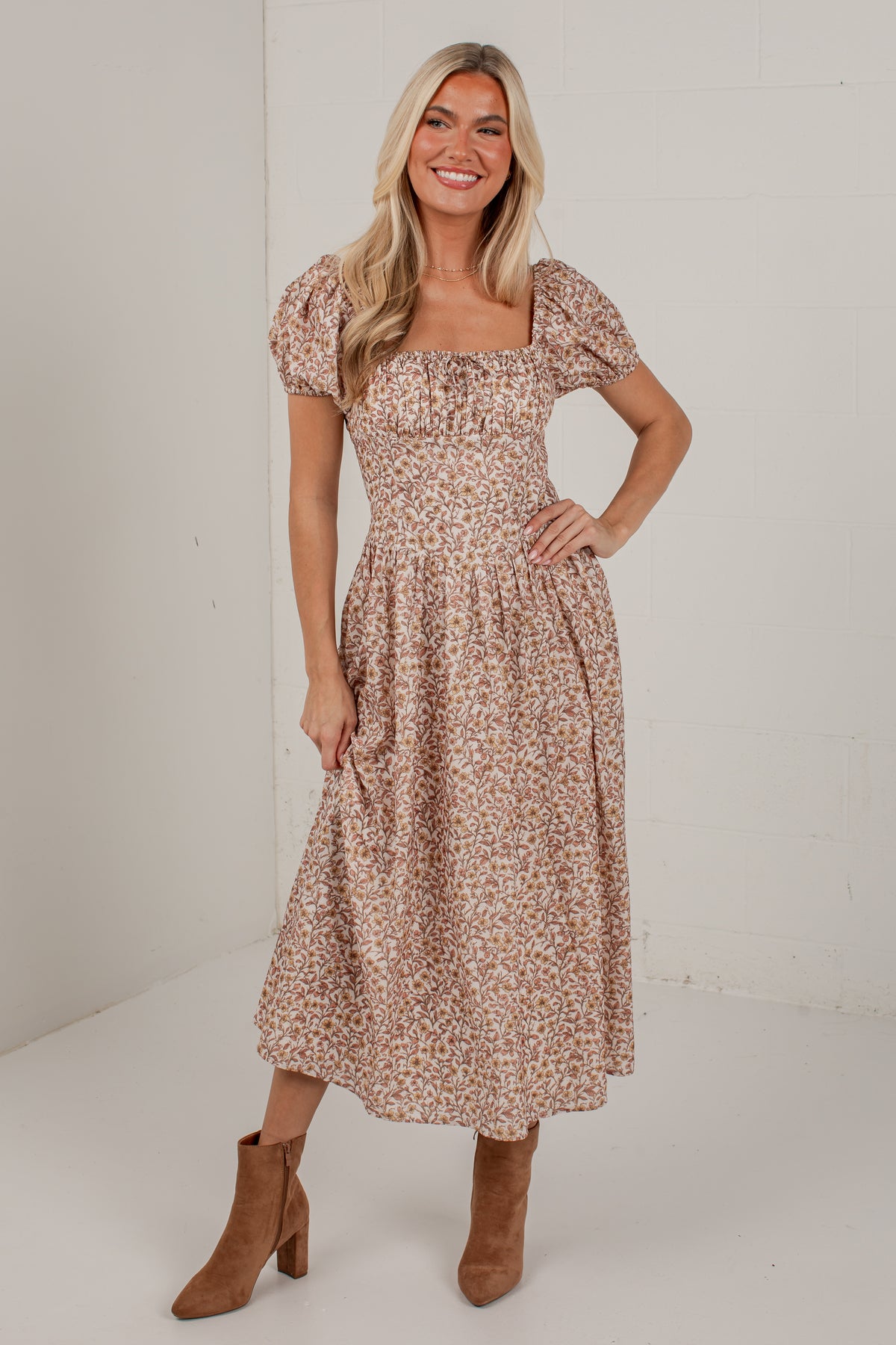 A woman modeling a midi-length puff-sleeve dress with a small-scale floral print in beige, brown, and muted orange tones, paired with brown ankle boots, set against a plain white studio background.