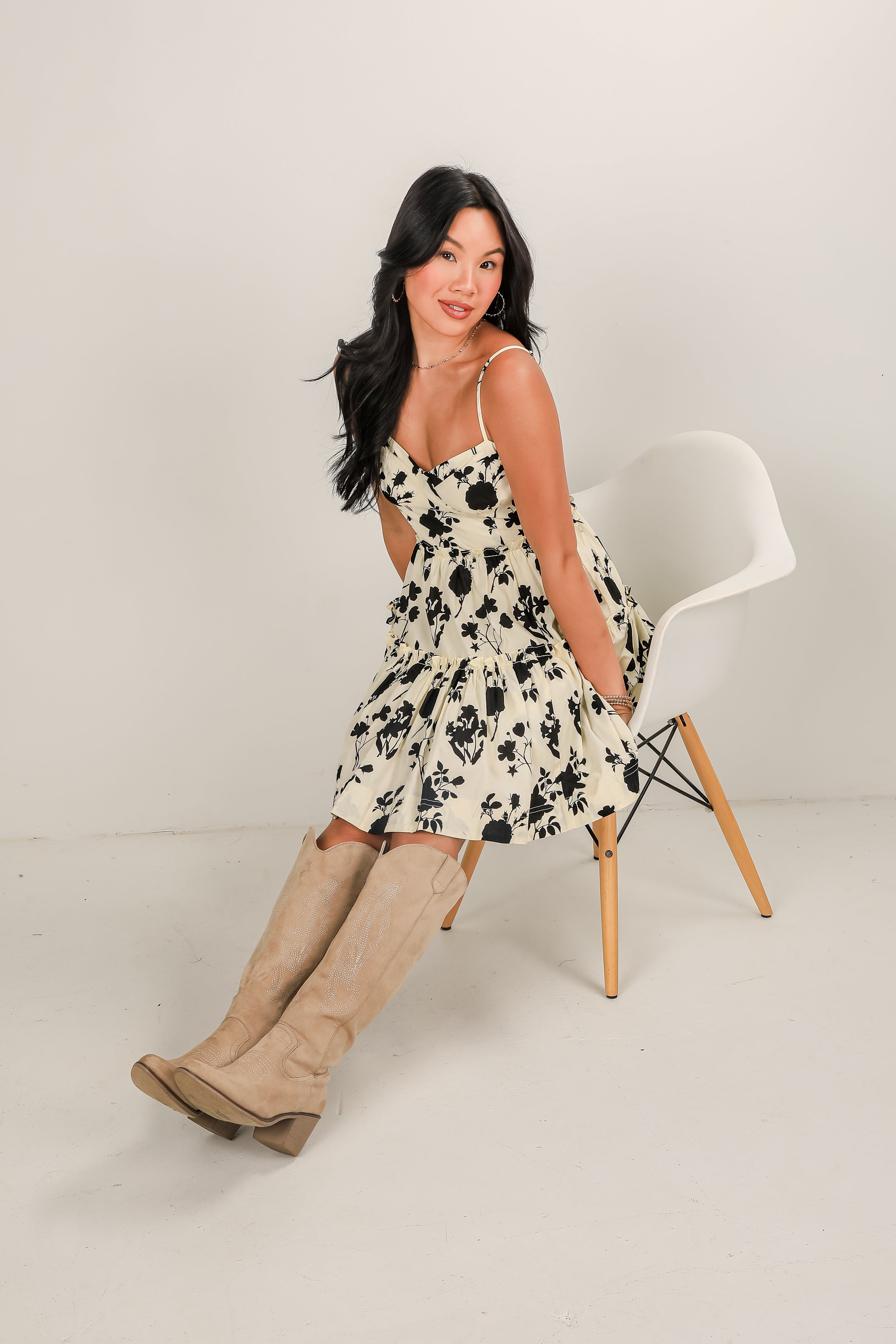 Woman in a floral dress and knee-high boots sitting on a chair against a plain background