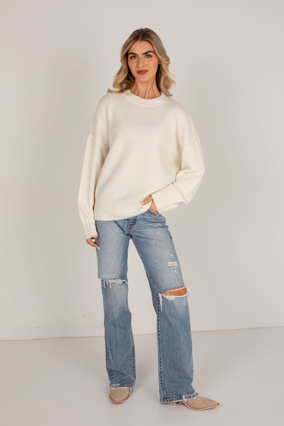 A woman modeling an oversized cream knit sweater and light-wash distressed flare jeans with tan pointed flats, standing against a plain off-white studio background.