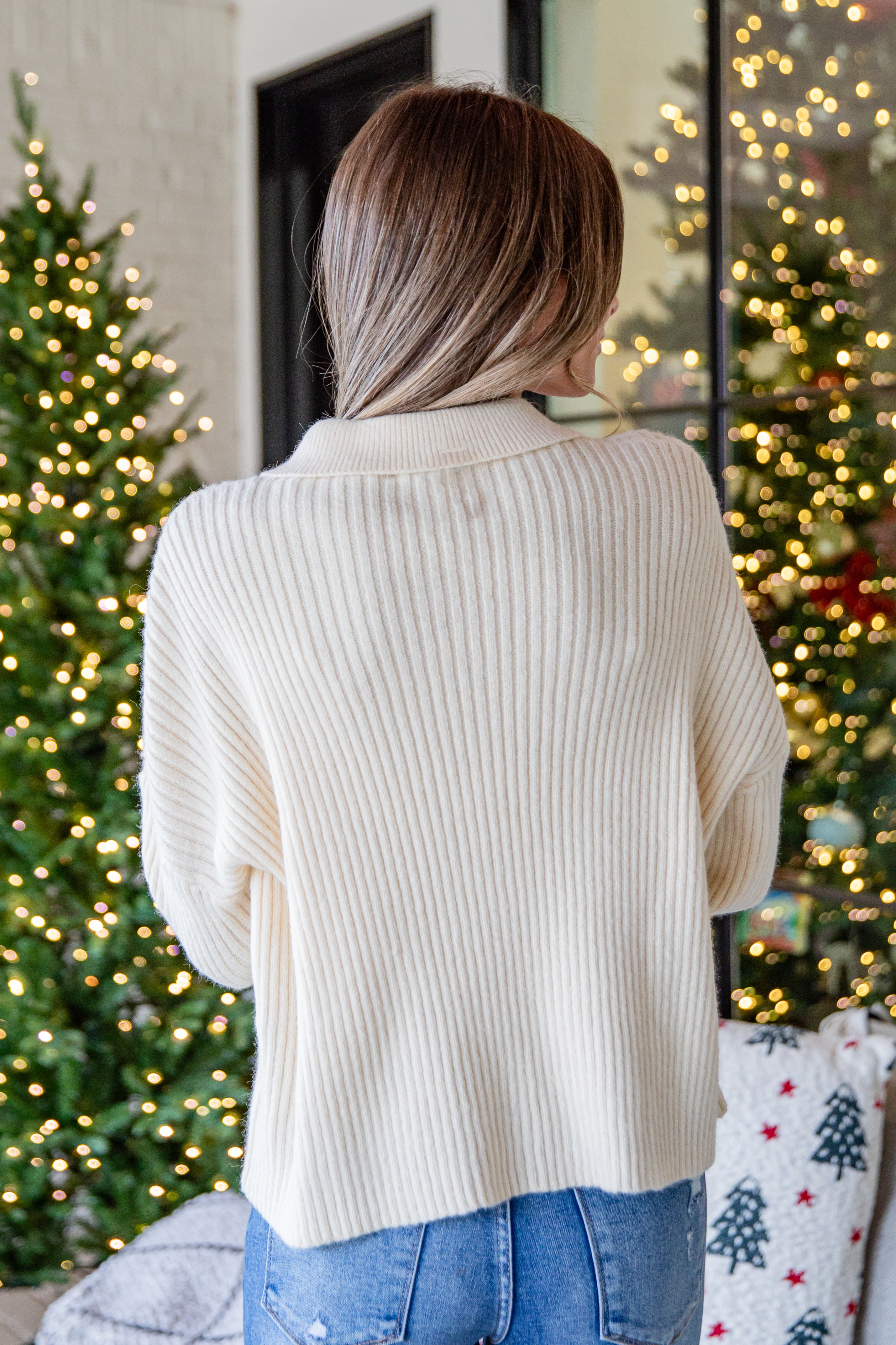 Woman wearing a beige sweater in front of Christmas trees