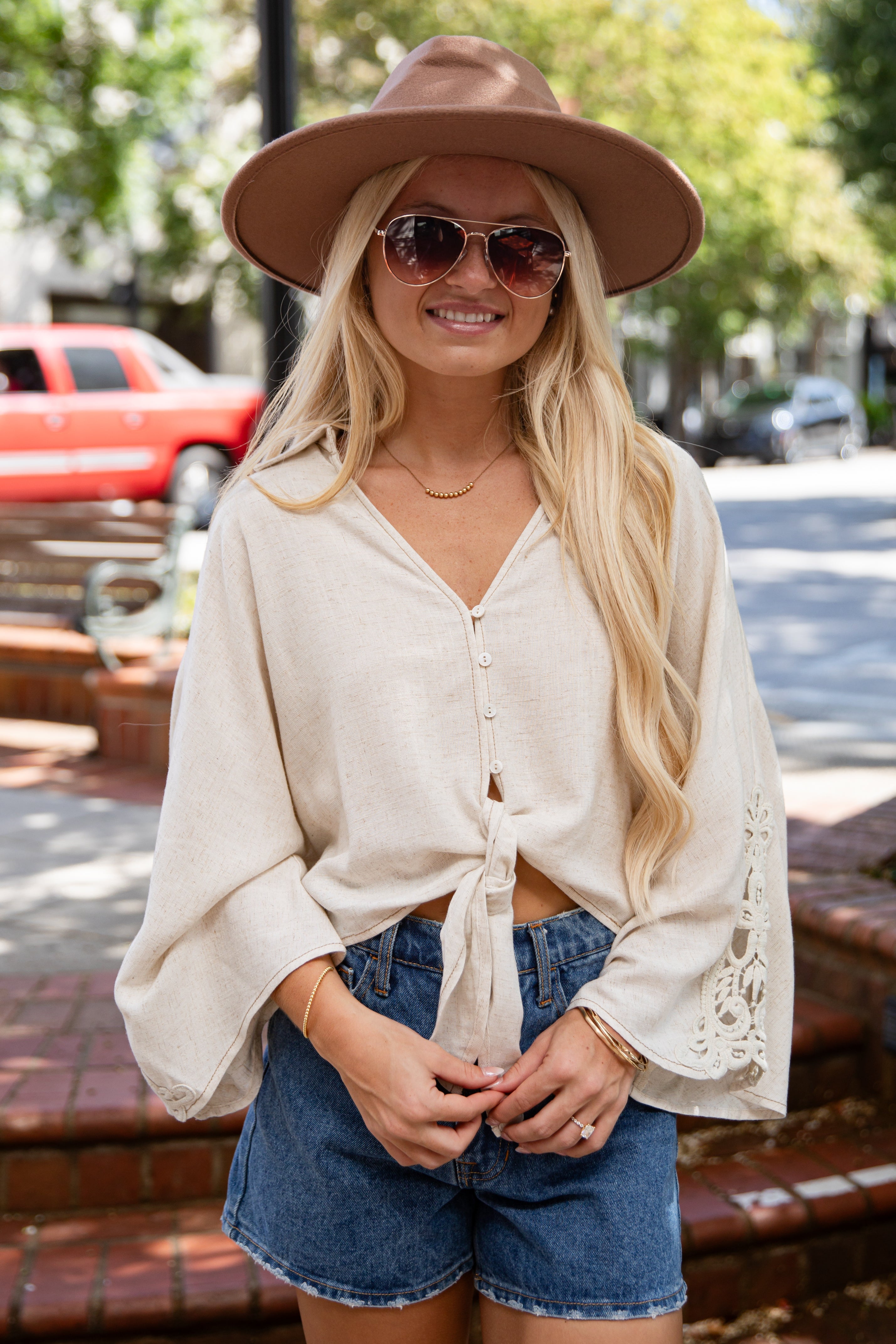 A woman wearing a cream-colored bohemian-style long-sleeve tie-front top, blue denim shorts, a brown wide-brim hat, and sunglasses, standing outdoors on a brick walkway with a blurred street background.