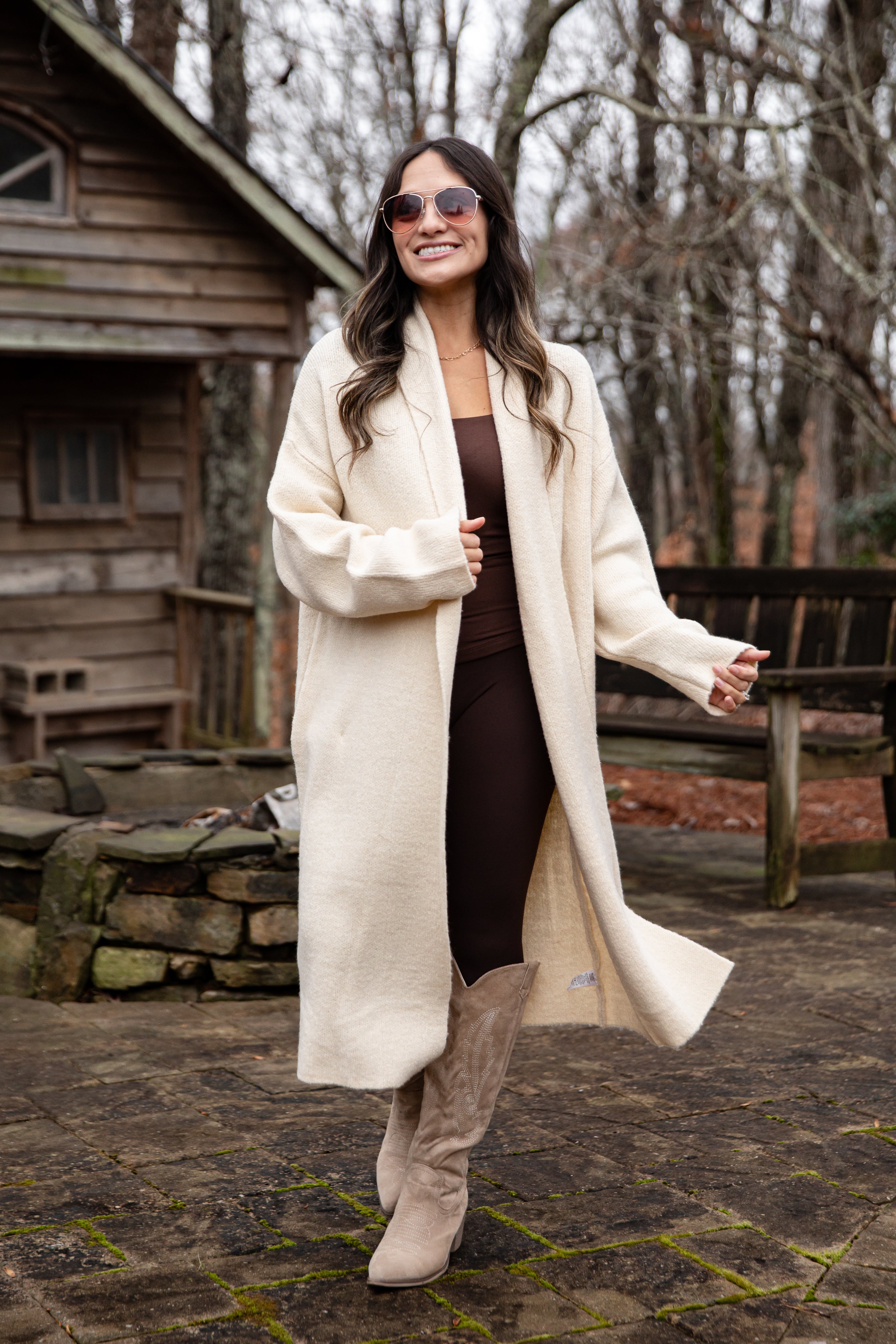 Woman in a long cream coat standing outdoors near a wooden cabin.