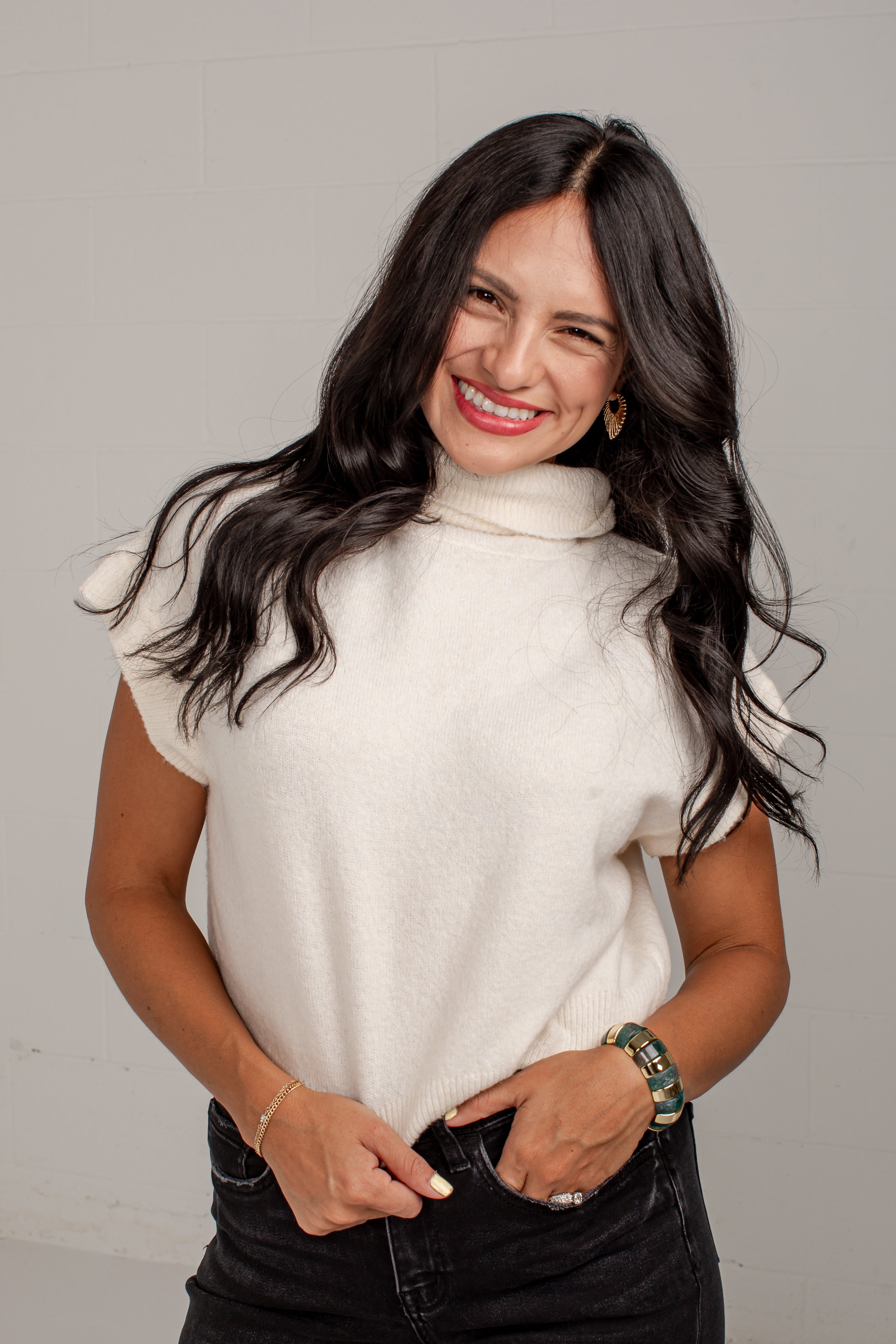 A smiling woman with long dark hair wearing a cream-colored short-sleeve mock neck sweater and black jeans, standing against a light gray wall.
