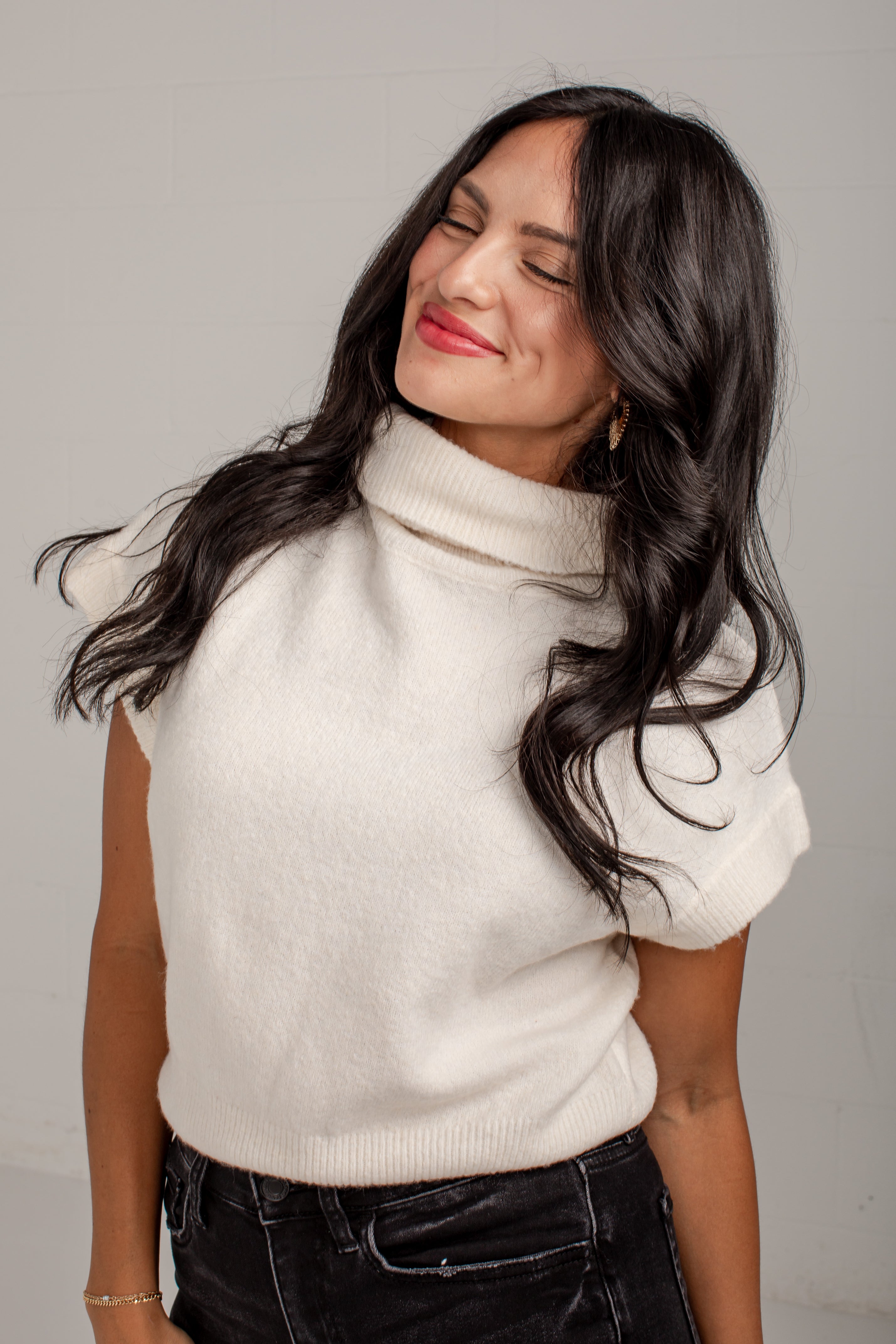 A woman with long, wavy black hair wearing a cream-colored short-sleeve mock neck knit sweater and black jeans, posing against a light gray background.