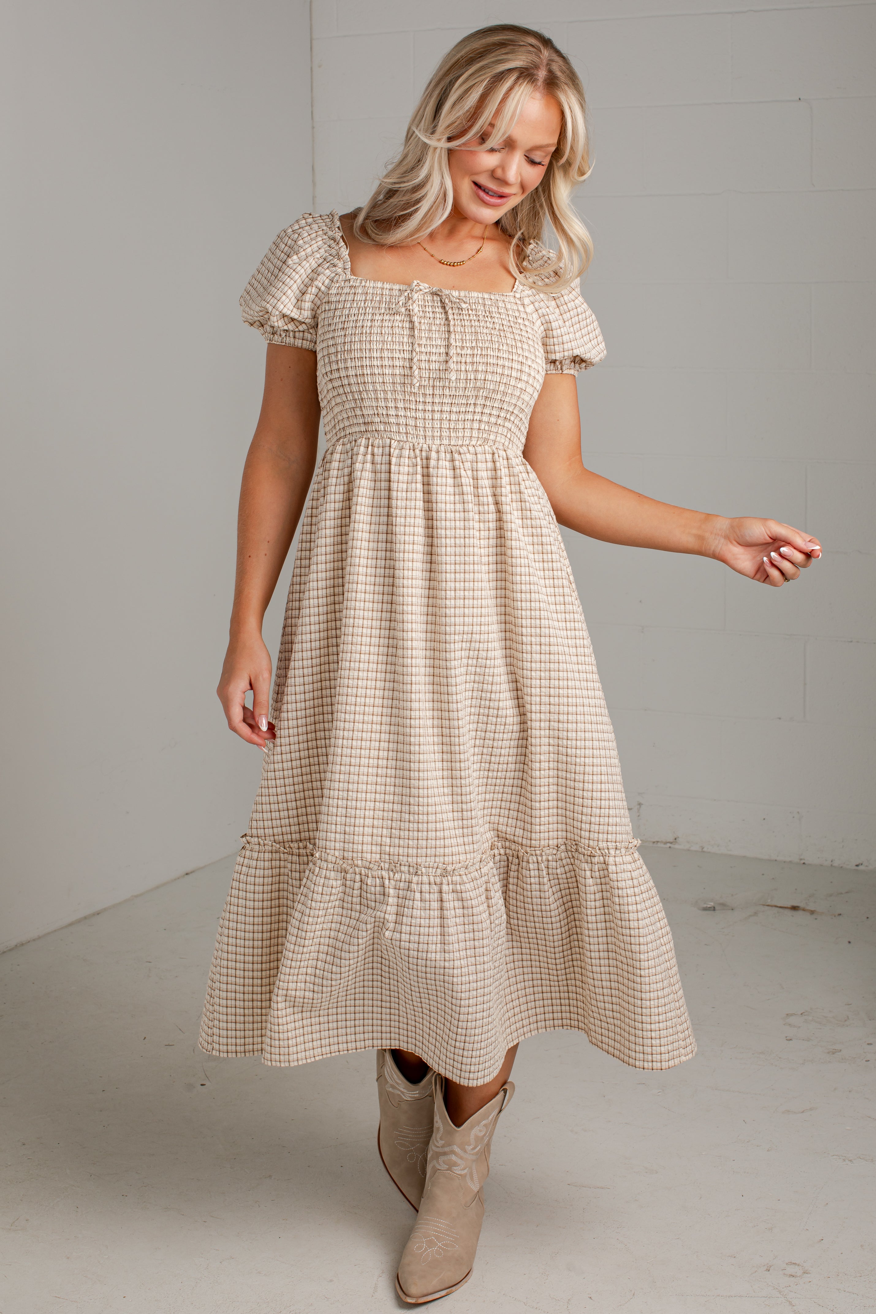 A woman modeling a beige and white gingham tiered midi dress with puff sleeves and a smocked bodice, paired with beige ankle boots, in a minimalist studio setting.