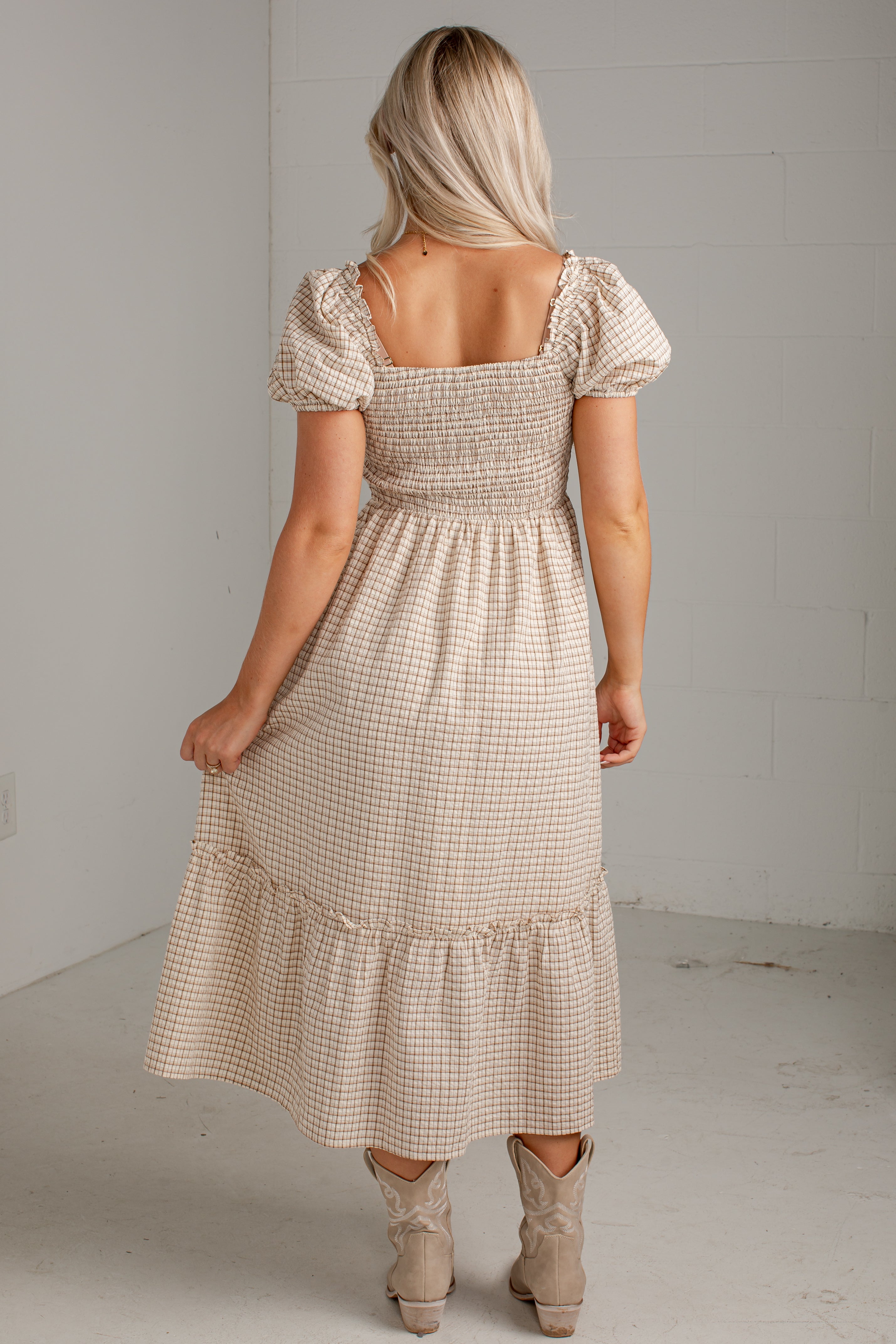 A woman modeling a beige and white gingham tiered midi dress with puff sleeves and a smocked bodice, paired with beige ankle boots, in a minimalist studio setting.