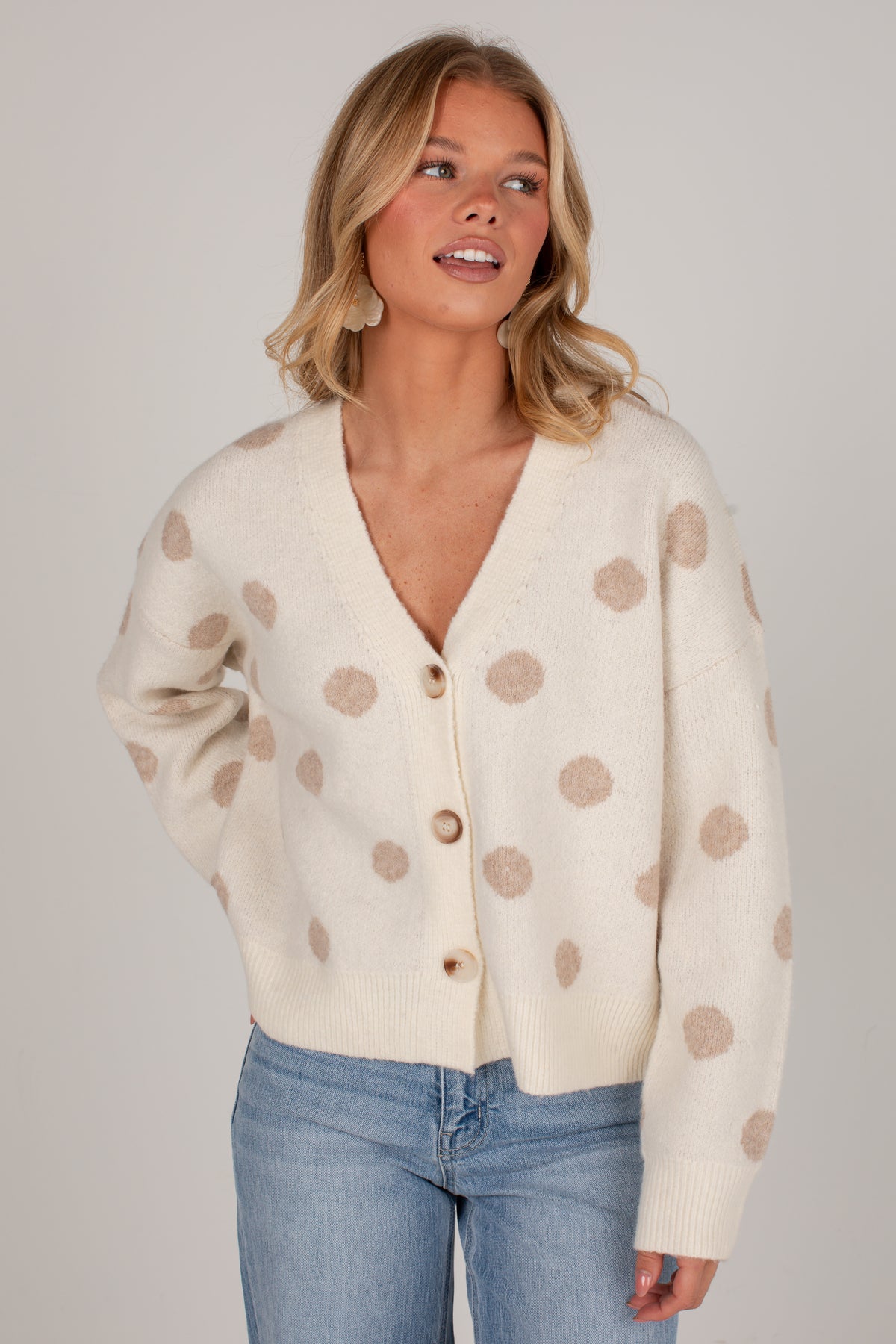 A woman wearing a cream-colored knit cardigan with beige polka dots and light blue jeans, photographed against a neutral light gray studio background.