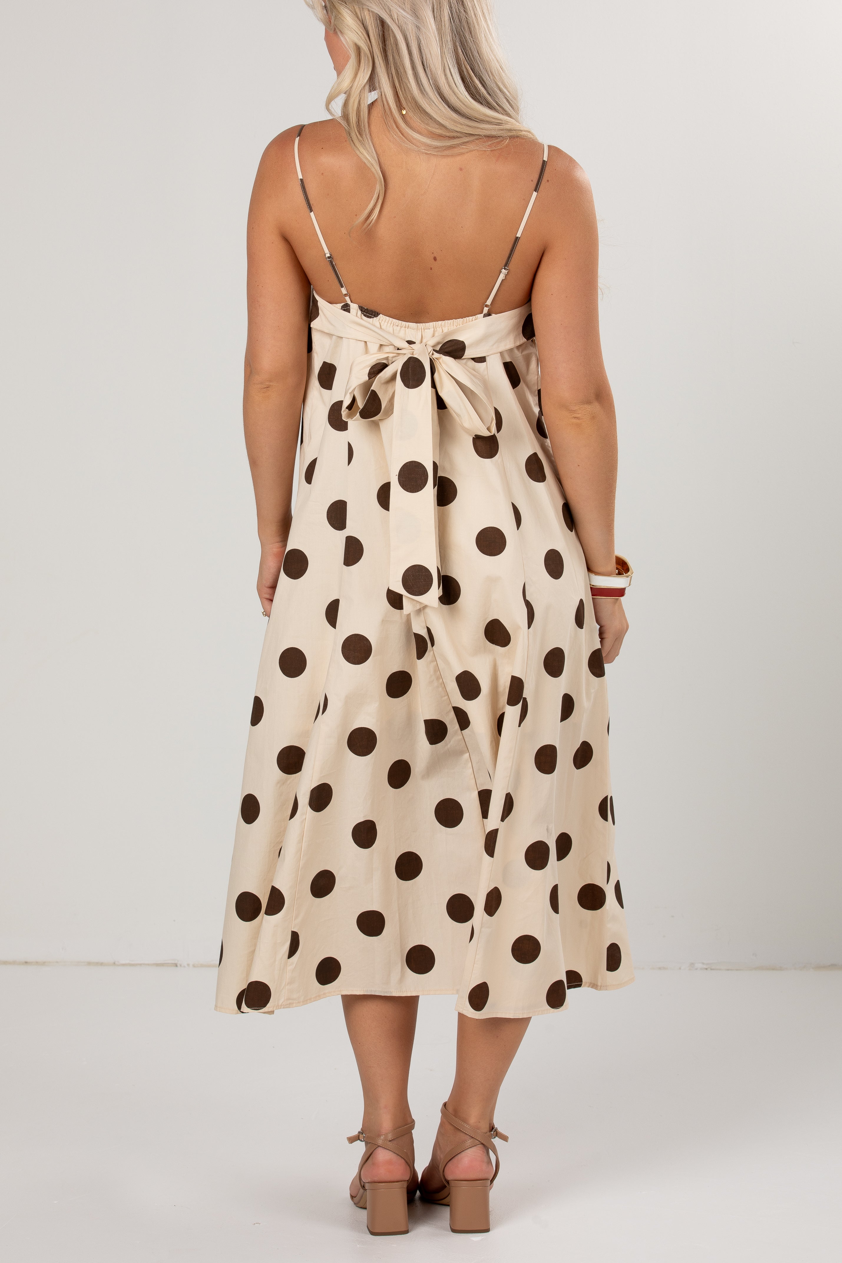A woman viewed from the back wearing a cream midi dress with dark brown polka dots and thin shoulder straps, paired with brown heeled sandals against a plain light gray studio background.