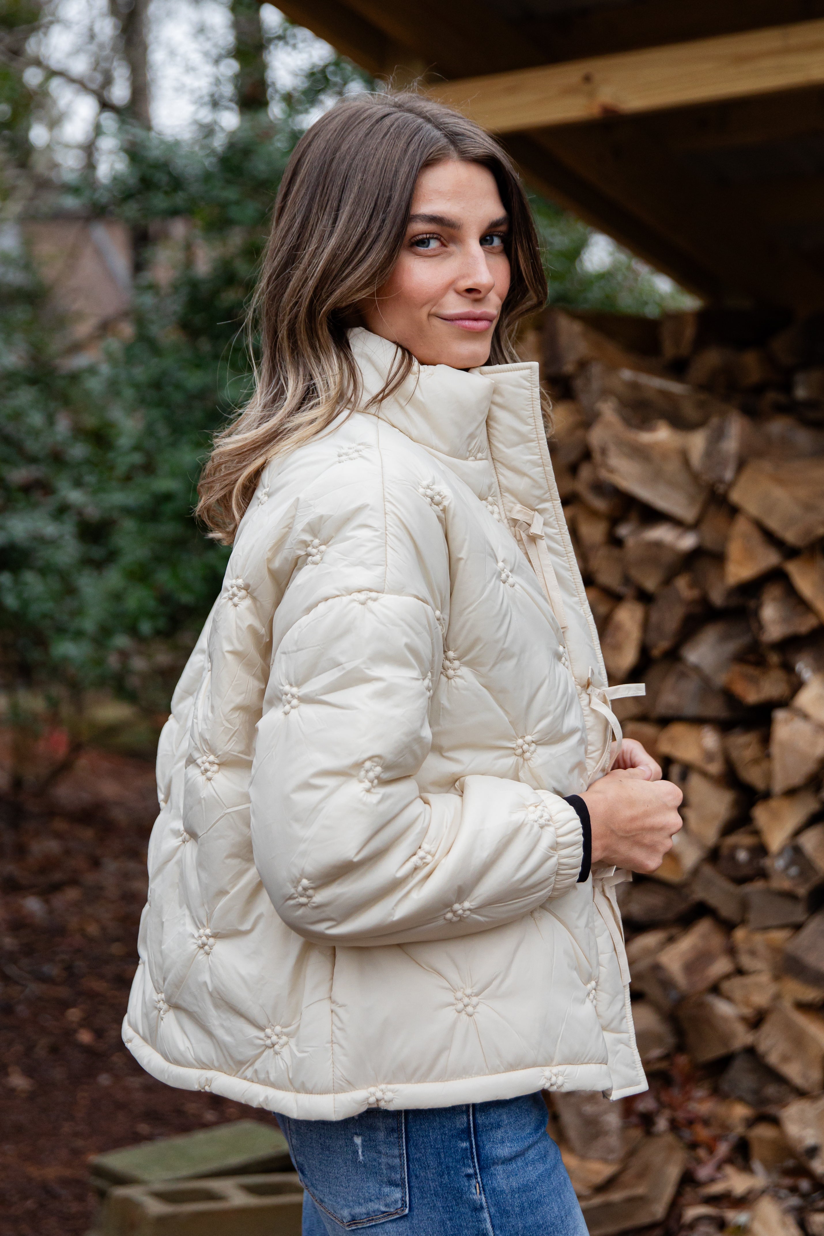 Woman wearing a white quilted jacket standing in front of stacked firewood.