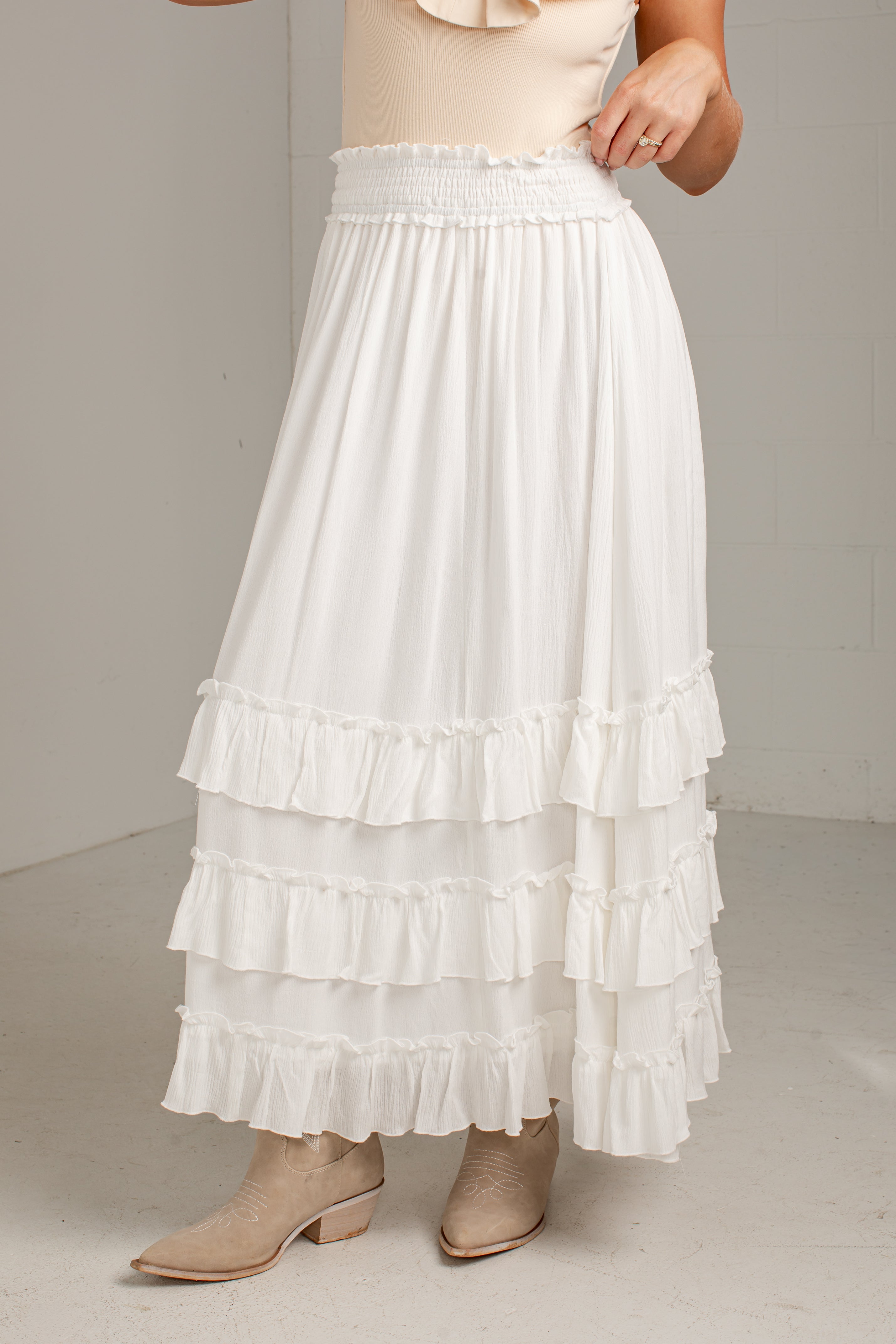 A white tiered maxi skirt with an elastic smocked waistband, shown on a model wearing beige ankle boots against a neutral studio background.
