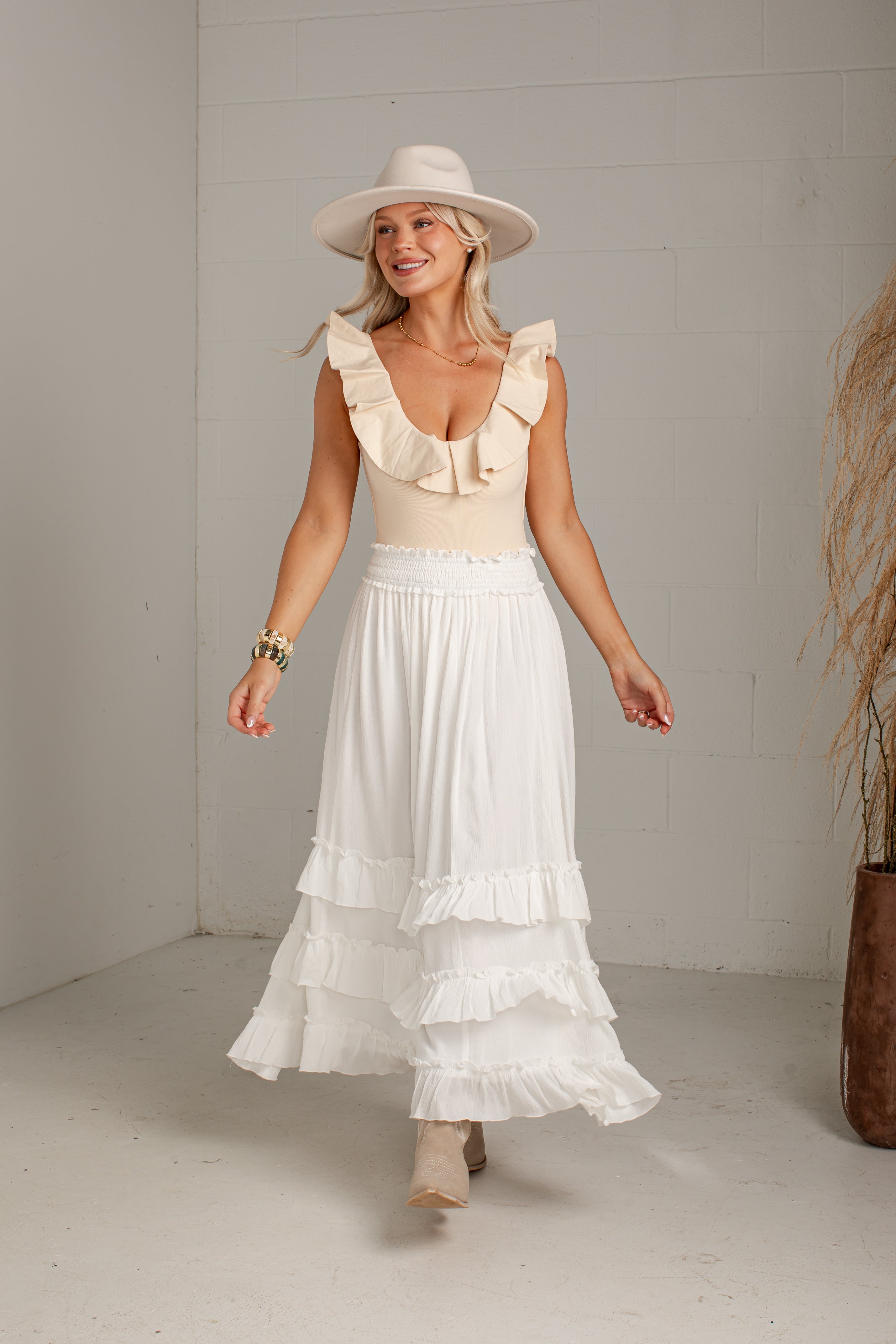A woman wearing a cream ruffled sleeveless top and a long white tiered skirt, paired with a wide-brimmed cream hat and beige boots, standing in a bright studio with a white wall.