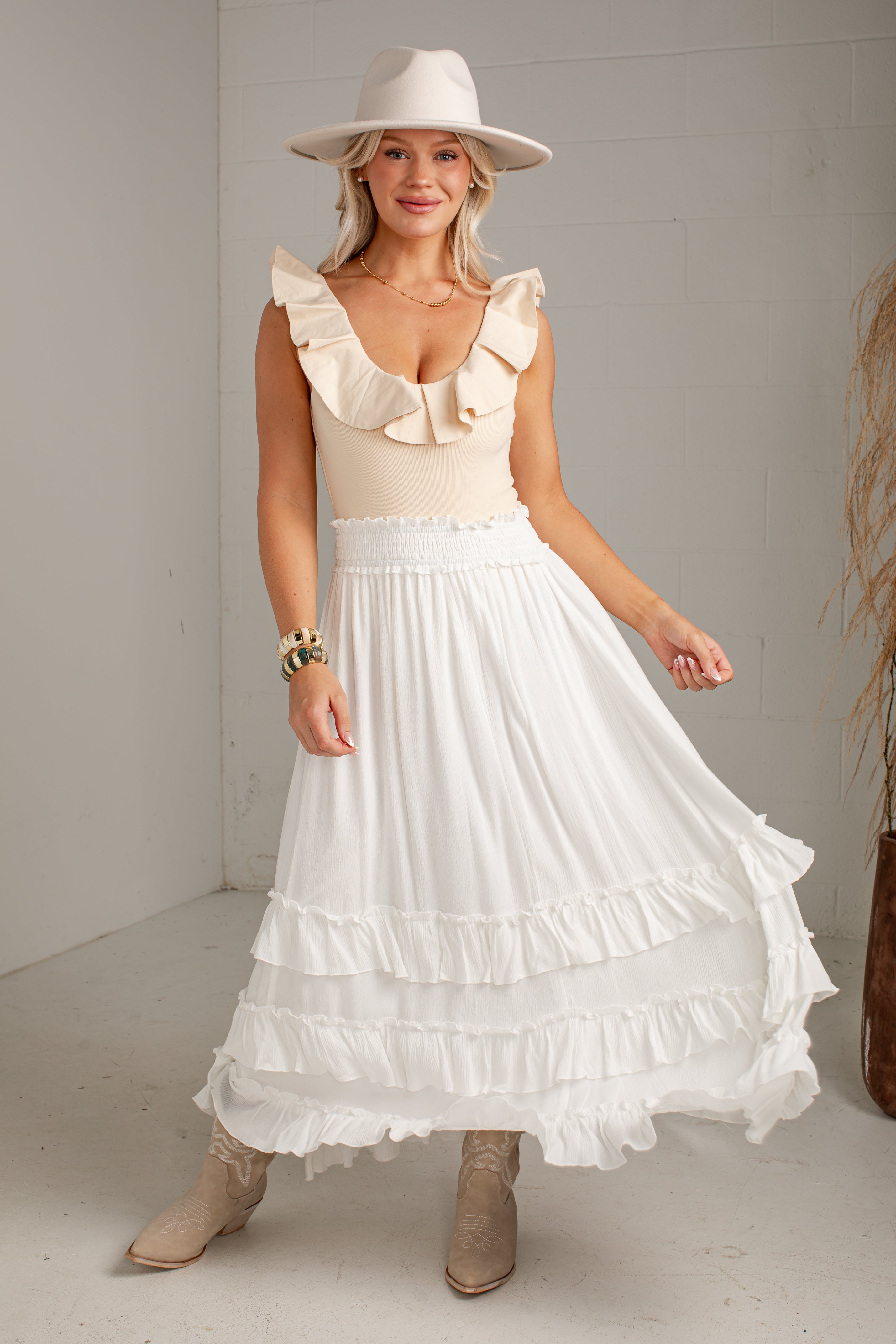 A woman modeling a bohemian-style white tiered maxi skirt with ruffle details, paired with a cream ruffled top, beige hat, and beige boots, standing in a minimalist white room.