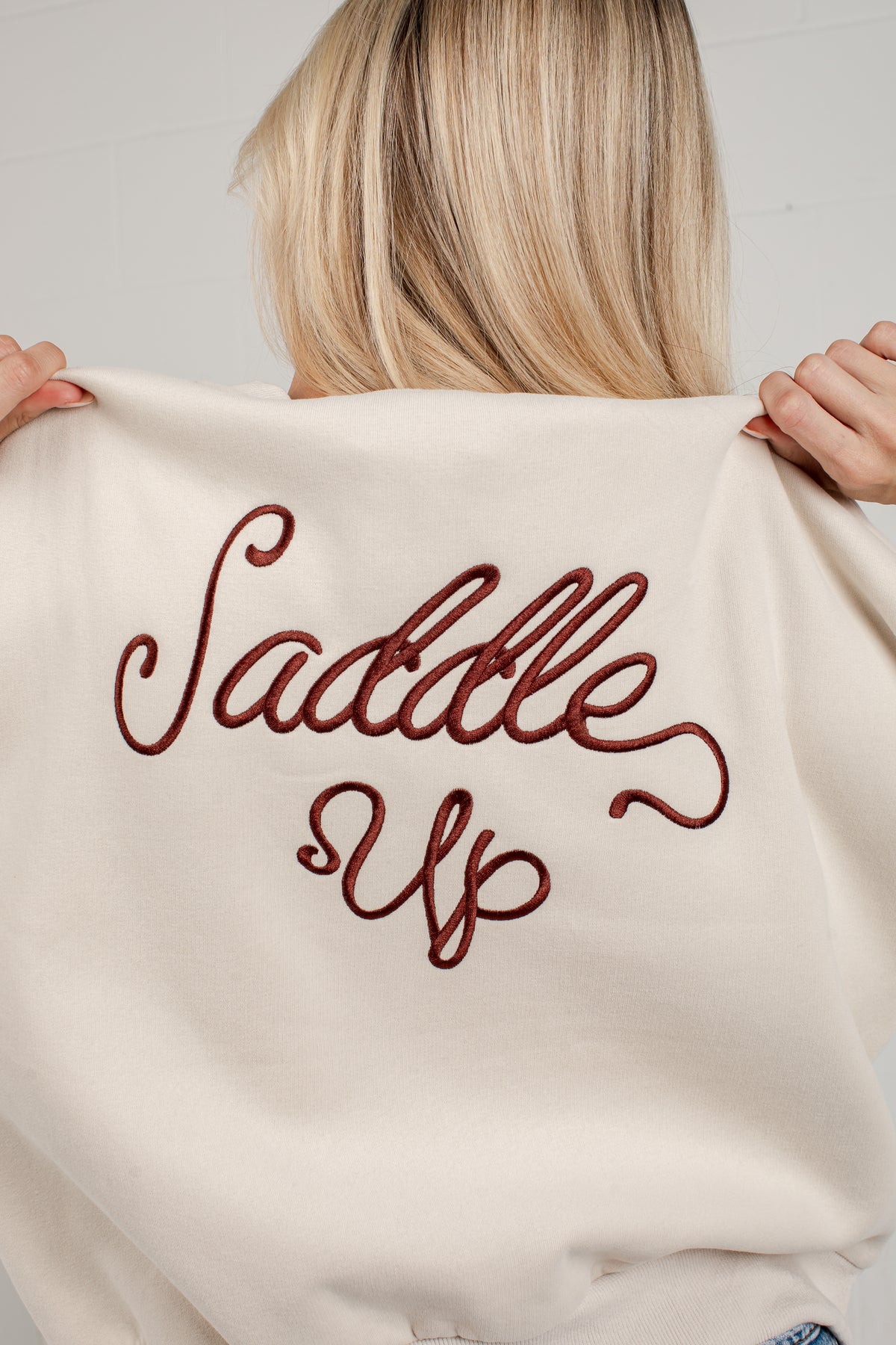 A person shown from behind holding up a cream-colored sweatshirt with the phrase &