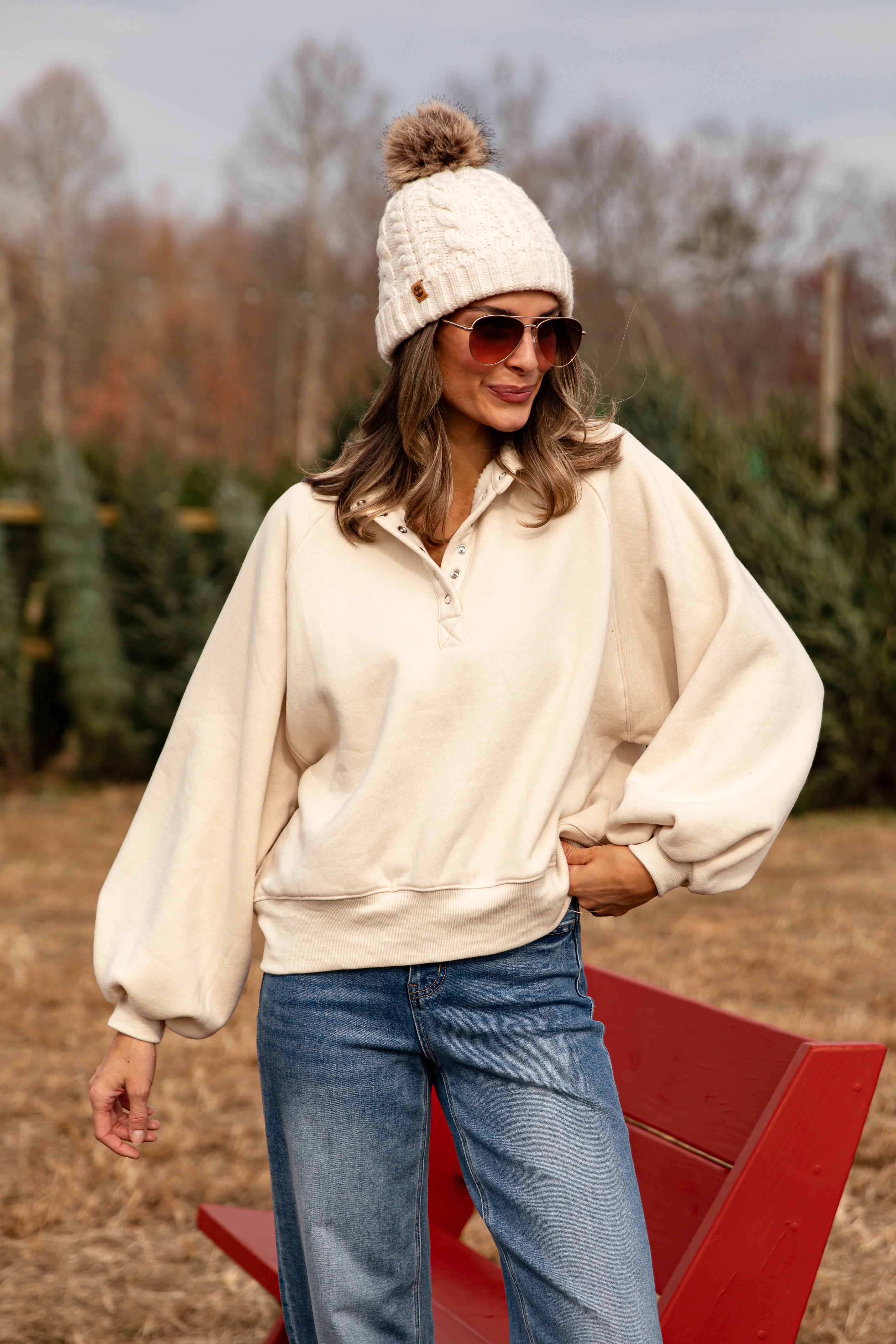 Woman wearing a cream sweater, blue jeans, and a white knit beanie with a pom-pom, standing in a field with trees.