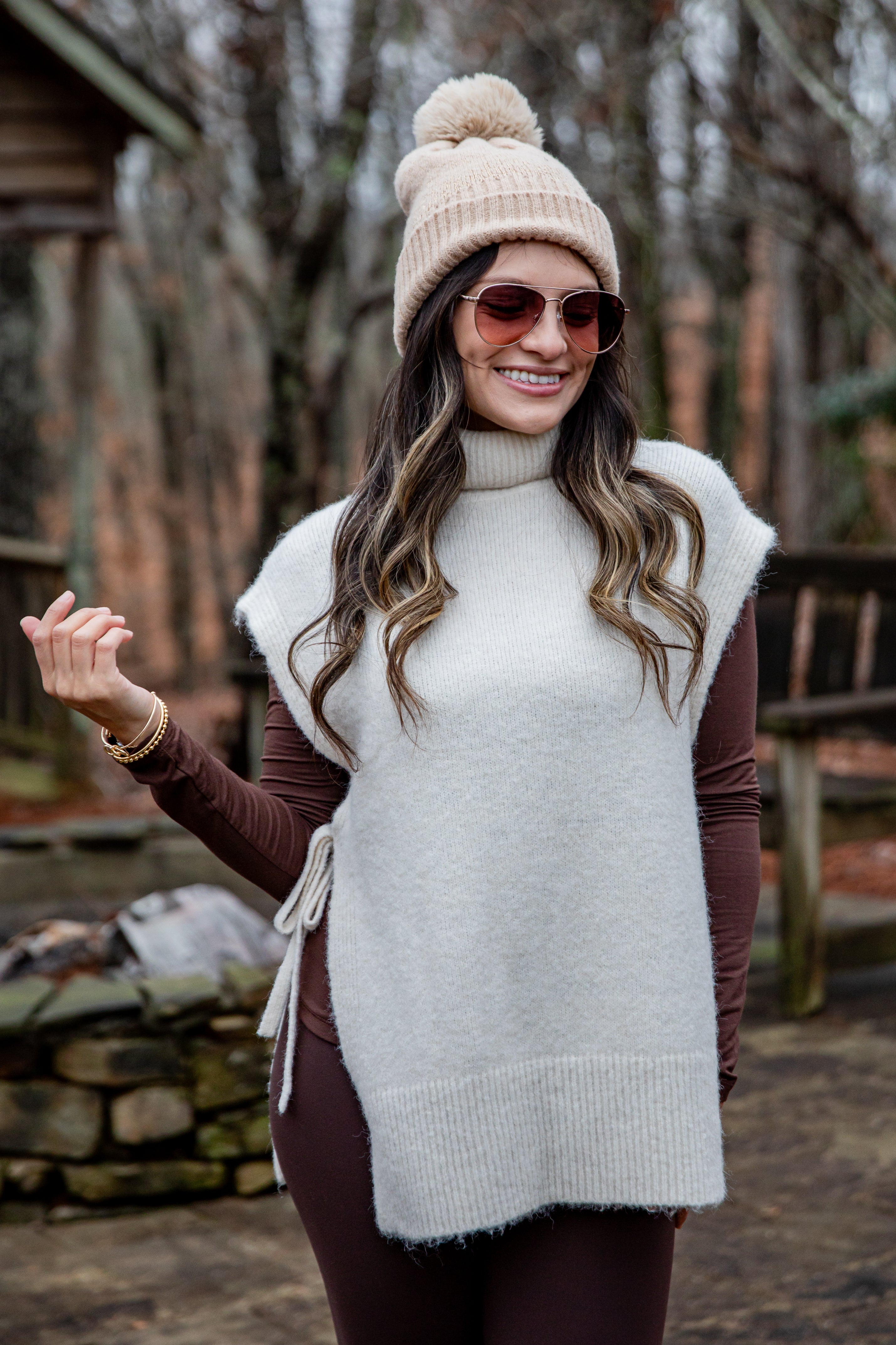 Woman wearing a white sweater vest, brown long-sleeve shirt, and beige knit hat outdoors.