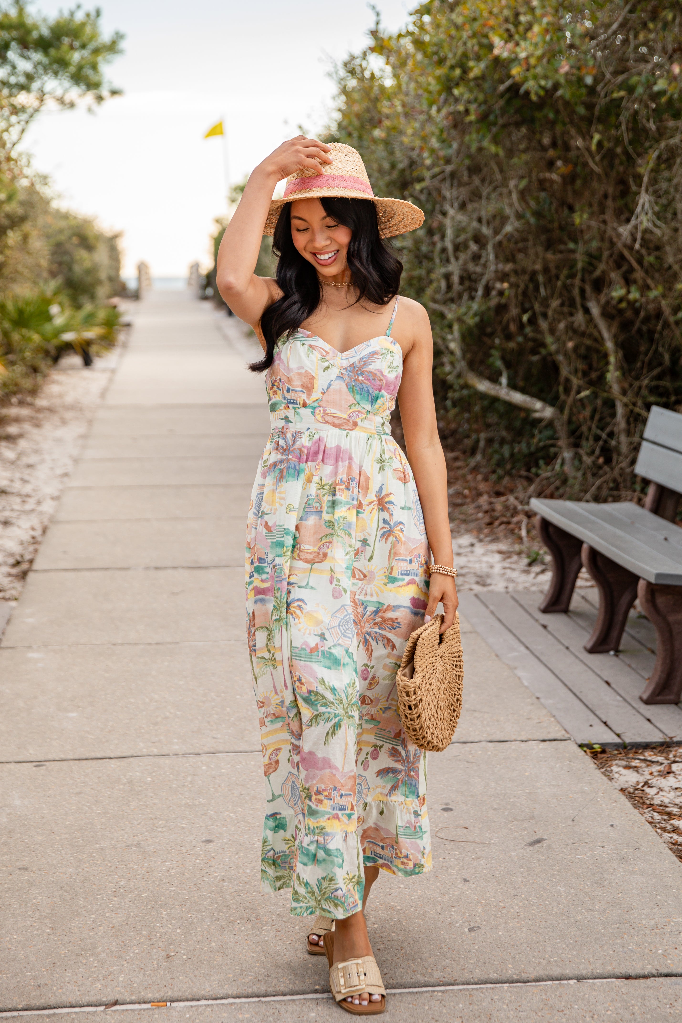 Woman in a floral dress and pink hat walking on a path with trees and a bench in the background.