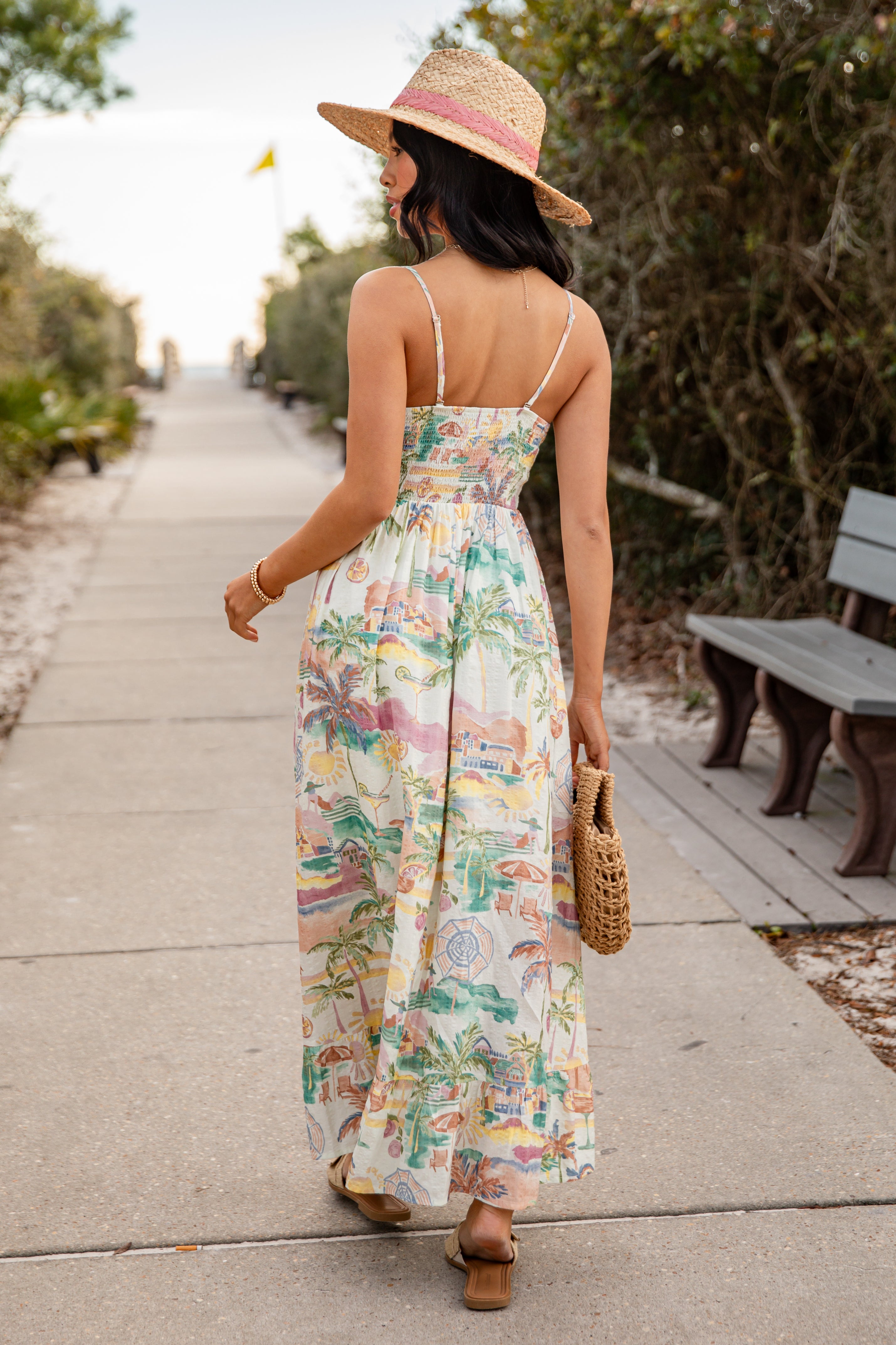 Woman in a floral dress and straw hat walking on a path with greenery.