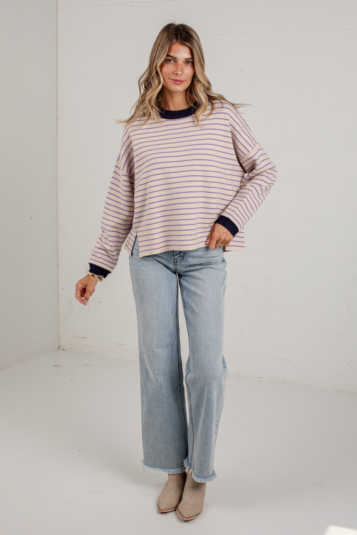 A woman modeling a long-sleeve striped knit top with navy blue trim, paired with light-wash wide-leg jeans and beige ankle boots, standing in a bright white studio corner.