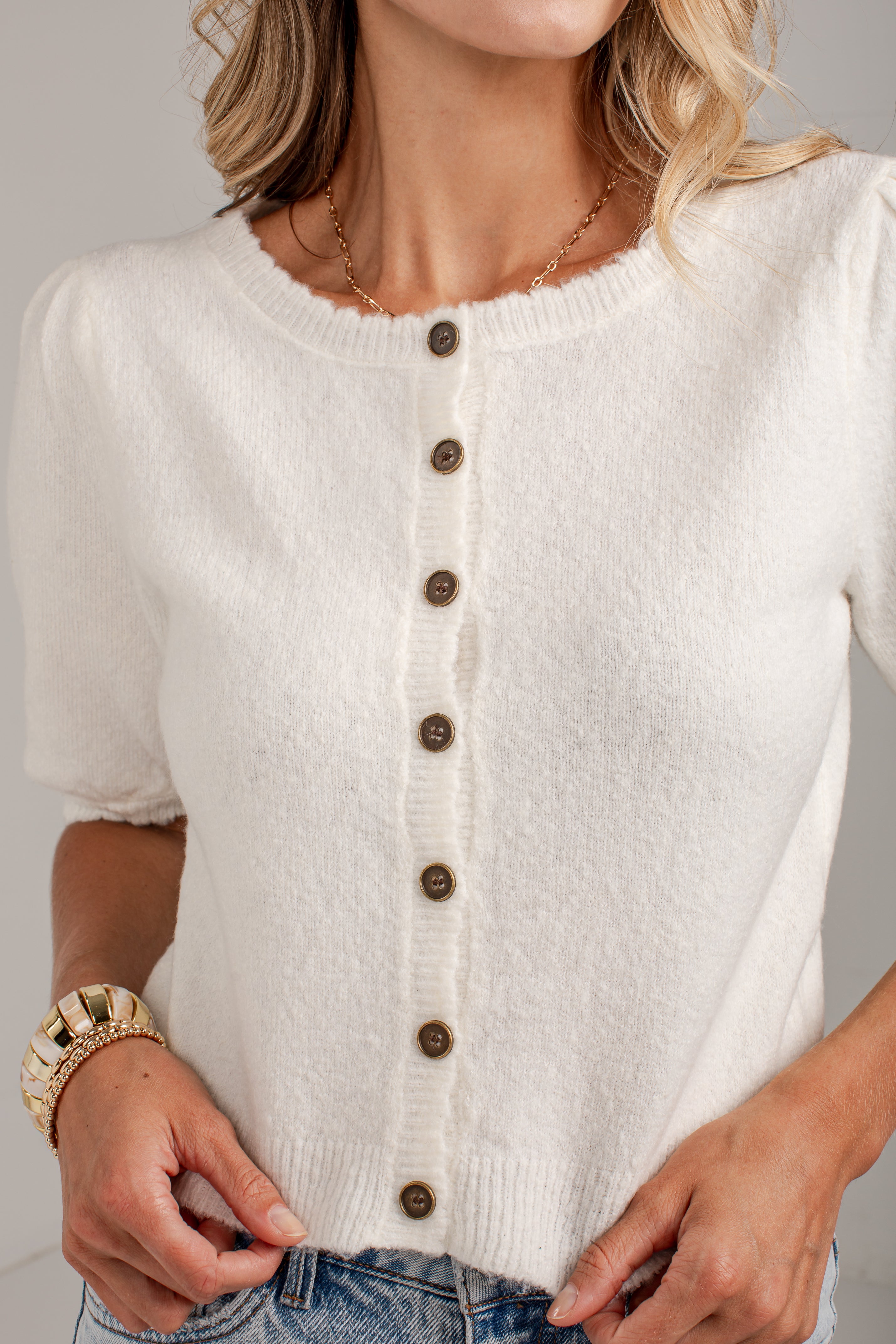 A close-up of a woman wearing a cream-colored short-sleeve knit cardigan with brown buttons, paired with blue jeans.