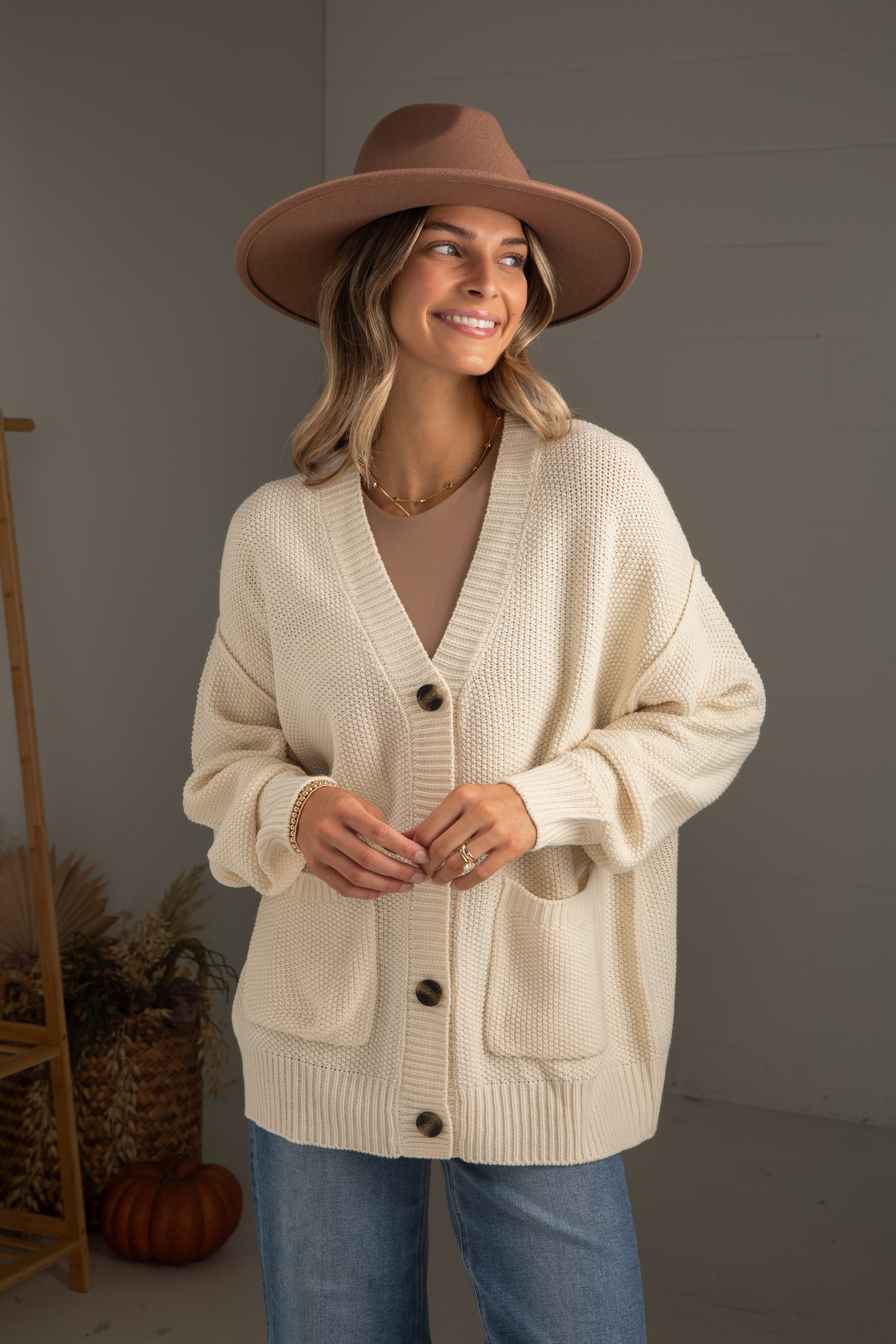 Woman wearing a beige cardigan, brown hat, and blue jeans indoors.