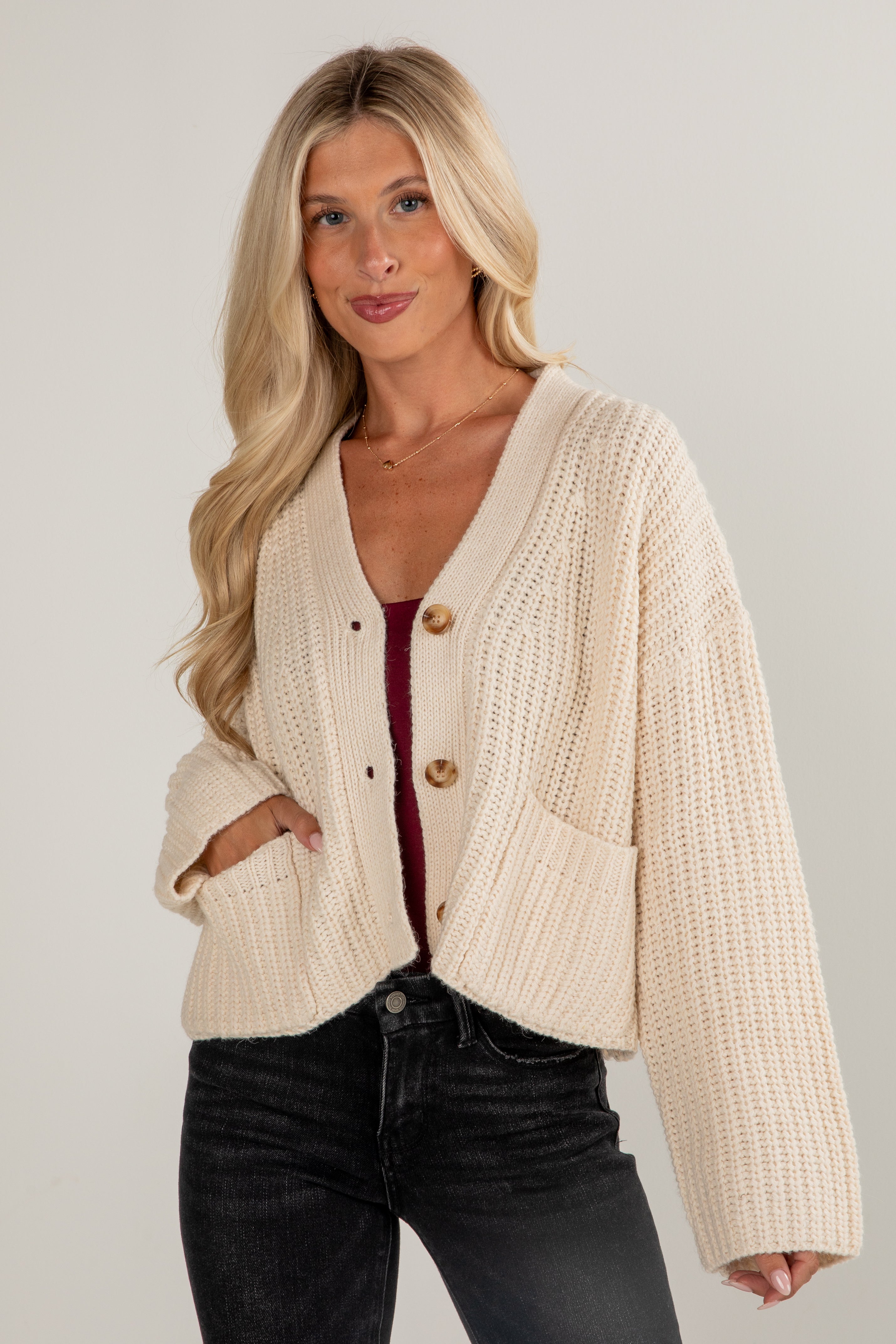 Woman wearing a beige knitted cardigan over a red top and black pants on a plain background