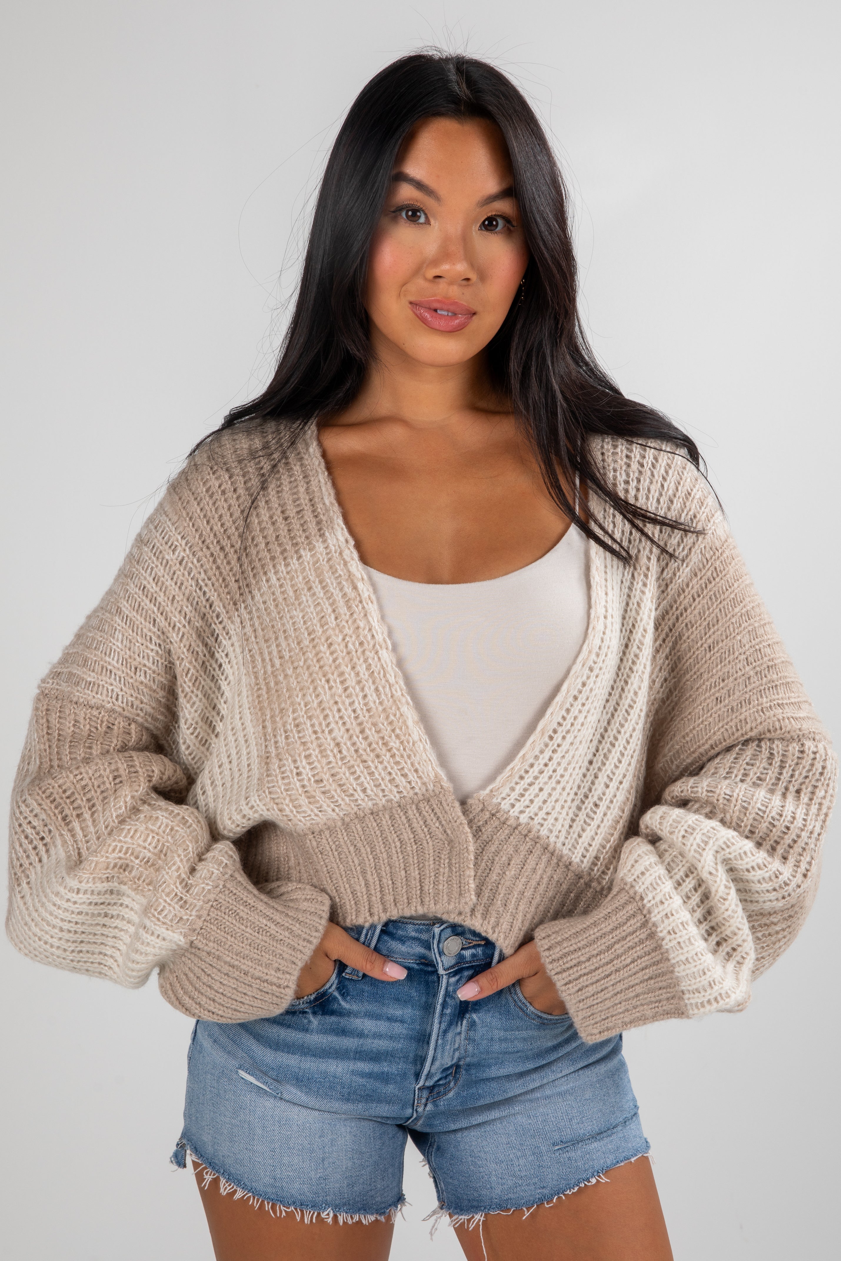 Woman wearing a beige knitted cardigan over a white top and denim shorts on a plain background