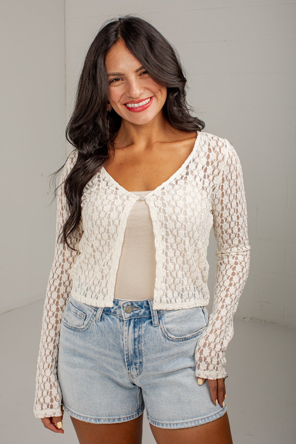 A woman modeling a cream-colored, long-sleeve crochet knit cardigan over a beige top and light-wash denim shorts, standing against a neutral light gray studio background.