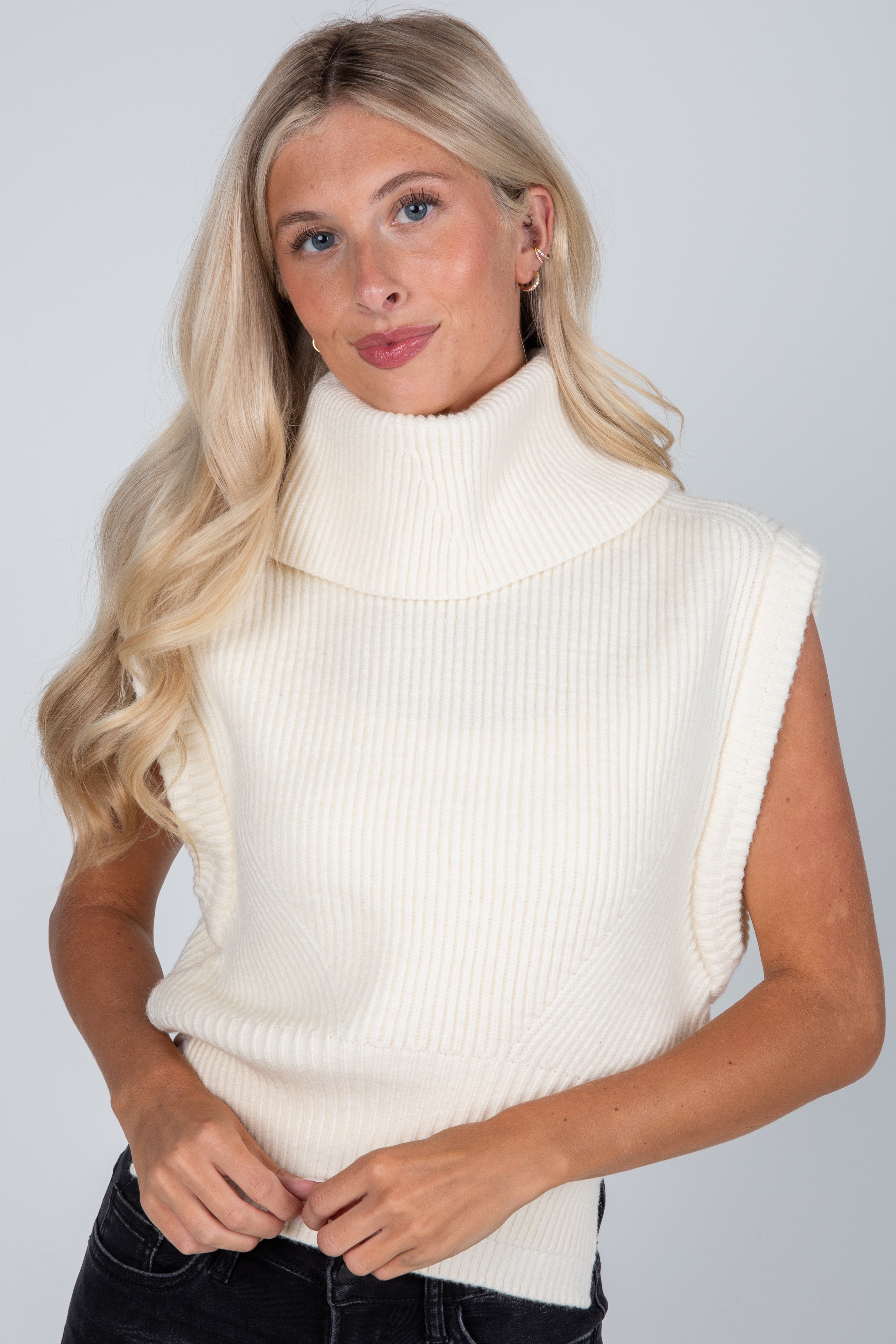 Woman wearing a cream sleeveless ribbed knit sweater against a plain background