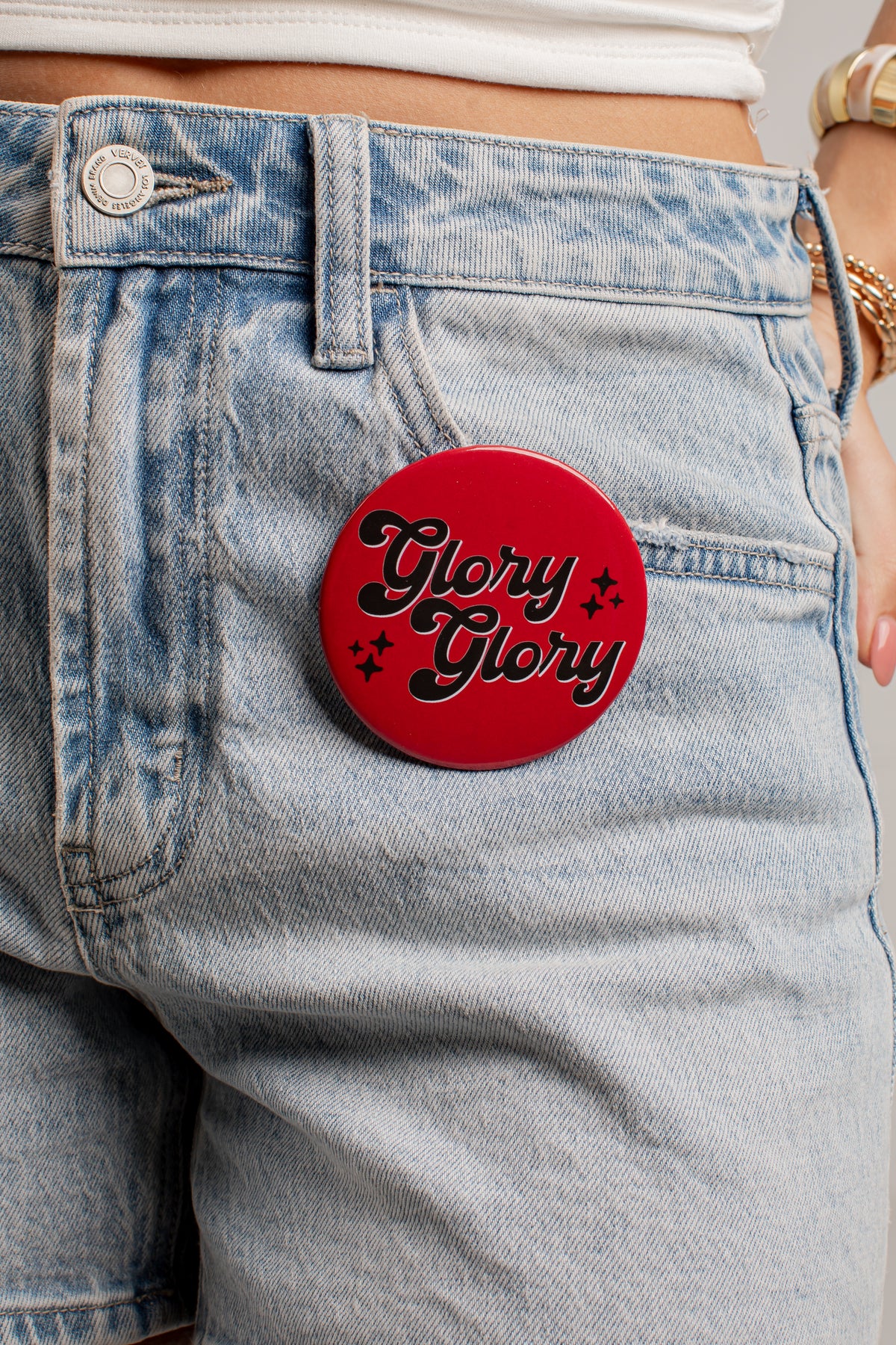 A close-up of a person wearing light-wash denim shorts and a white cropped top, with a round red pin on the back of the shorts that reads &